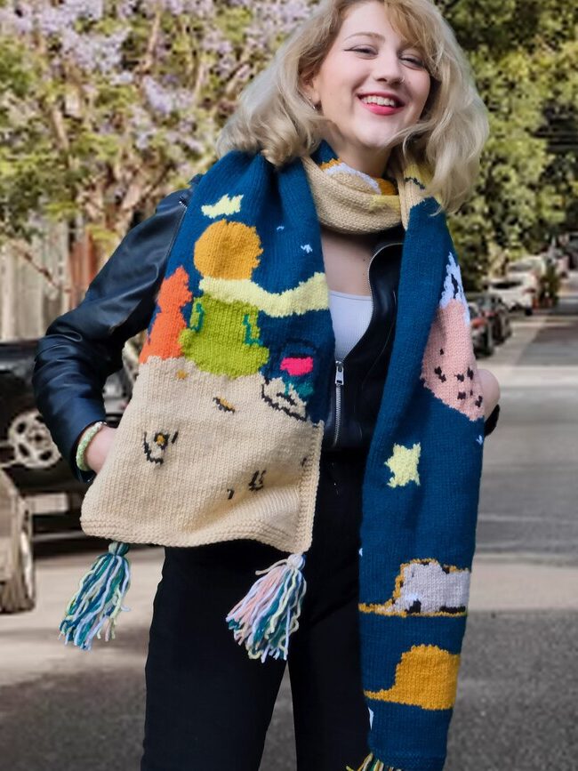 Person standing on a street wearing a colorful scarf with various designs and a black jacket, smiling, with parked cars and trees lining the road.
