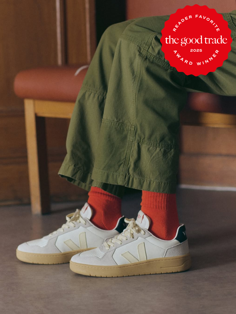A person wearing olive green pants, red socks, and cream-colored sustainable sneakers from Veja sits on a red cushioned chair. A red "The Good Trade 2025 Reader Favorite Award Winner" badge is in the top right corner.