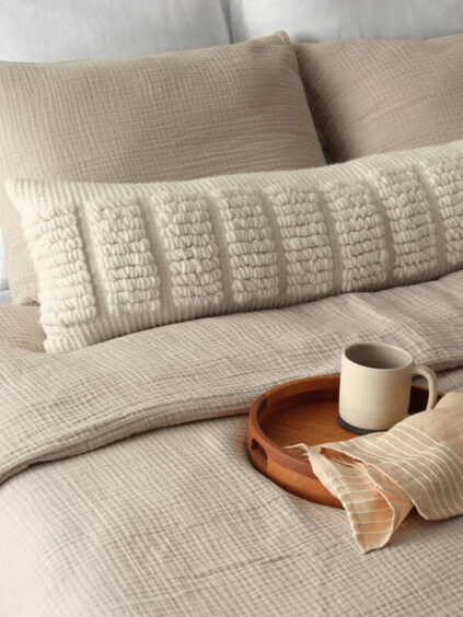 A neatly made bed with textured beige bedding, a decorative long pillow, and a wooden tray holding a beige mug and folded napkin.