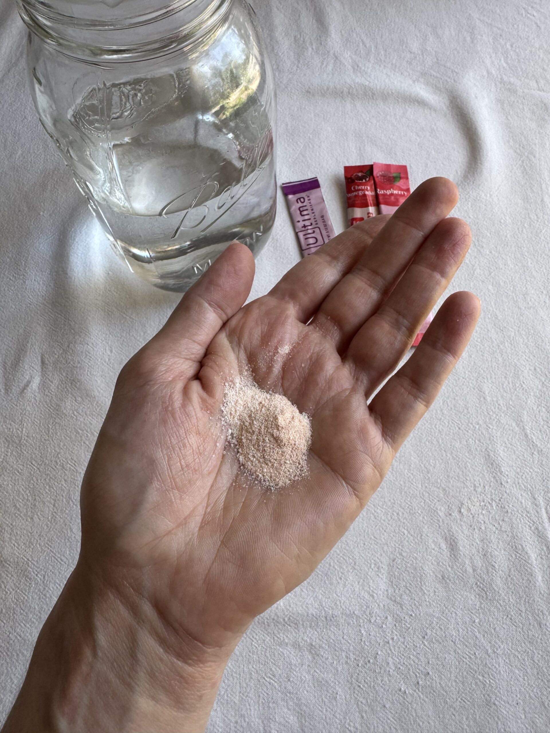 A hand holds a small pile of pink powder, with a glass jar of clear liquid and two small packets on a white surface in the background.