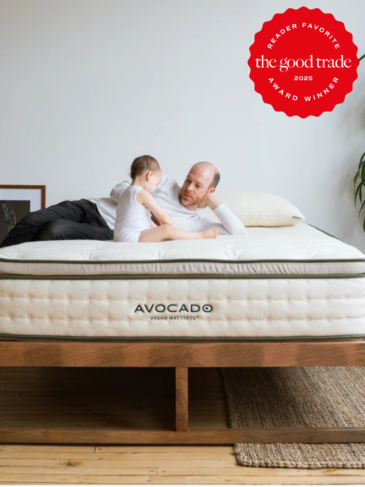 An adult and child sit on an Avocado Vegan Mattress in a minimalist bedroom. A red badge in the corner reads "The Good Trade 2025 Award Winner.