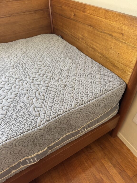 A neatly made bed with a patterned gray mattress and a wooden headboard and footboard on a wooden floor.