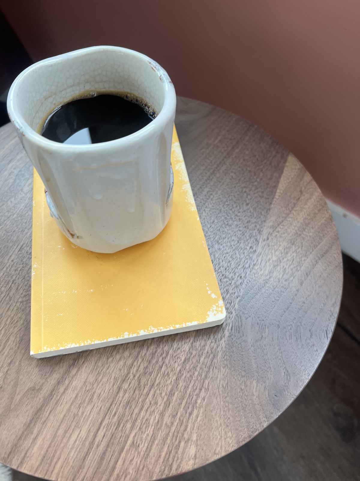 A white ceramic cup filled with black coffee sits on a worn orange book atop a round wooden table.