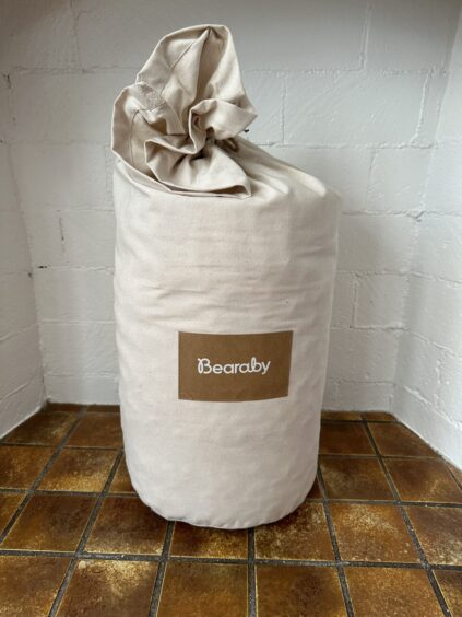 A rolled-up Bearaby weighted blanket in a beige fabric bag sits on a brown tiled floor against a white brick wall.