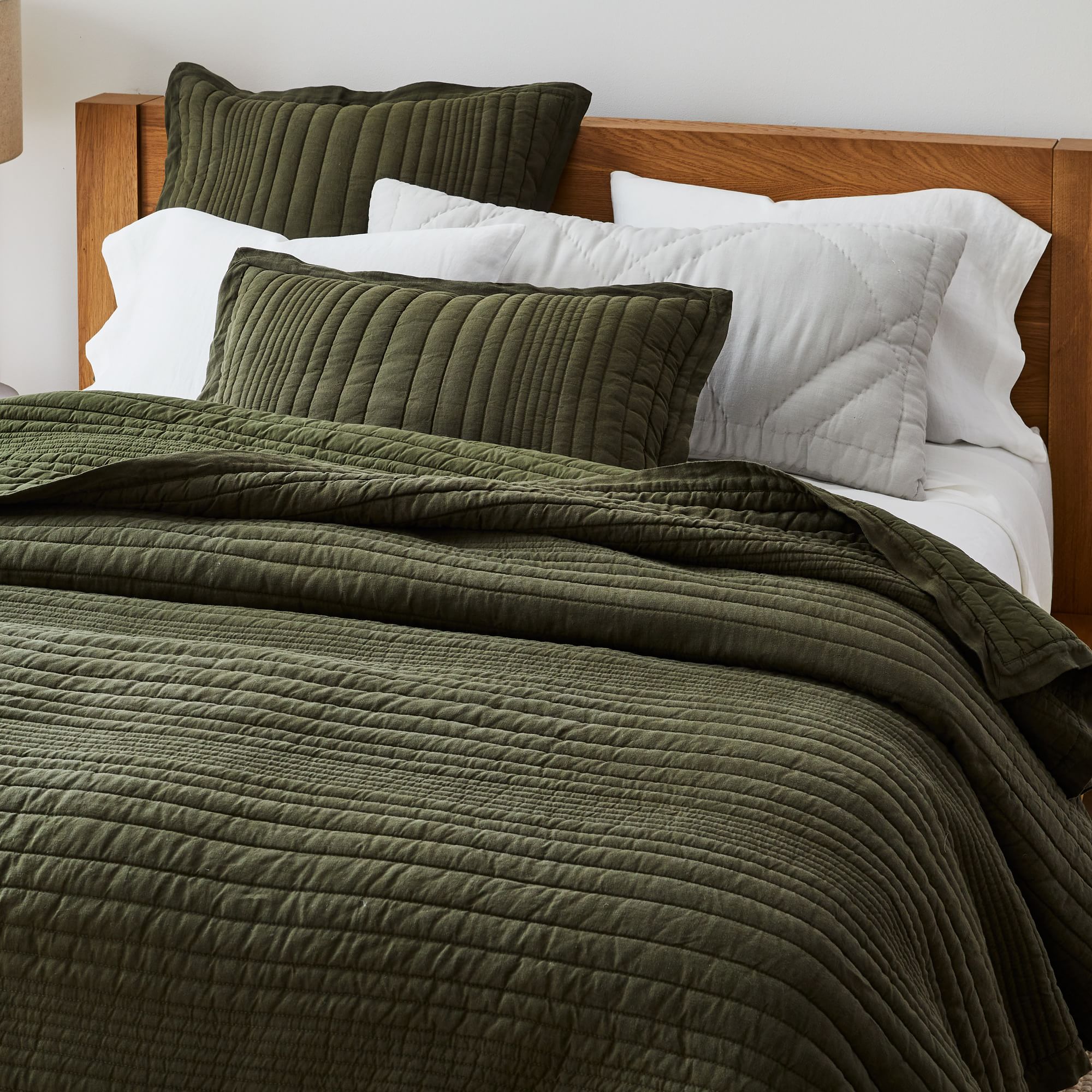 A neatly made bed with an olive green quilt and pillows. The bedding includes two dark green pillows and two white ones, all against a wooden headboard.