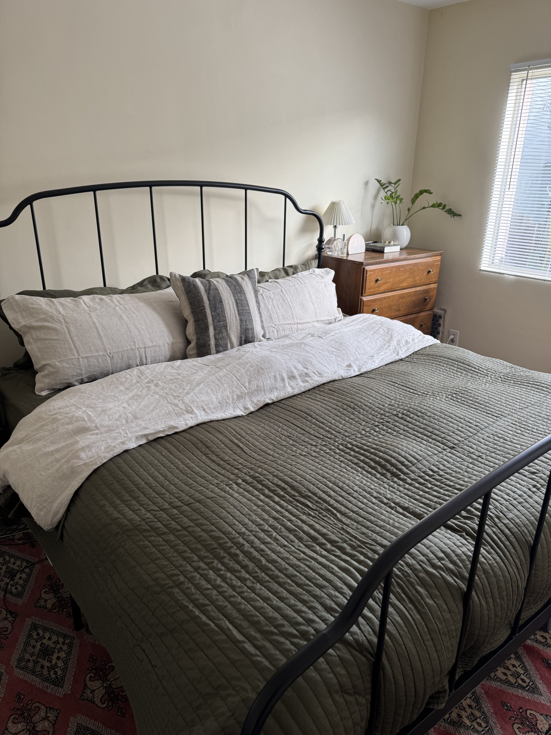 A neatly made bed with a black metal frame, green quilt and pillows next to a wooden dresser with a lamp and plant. Natural light enters through a window with blinds.