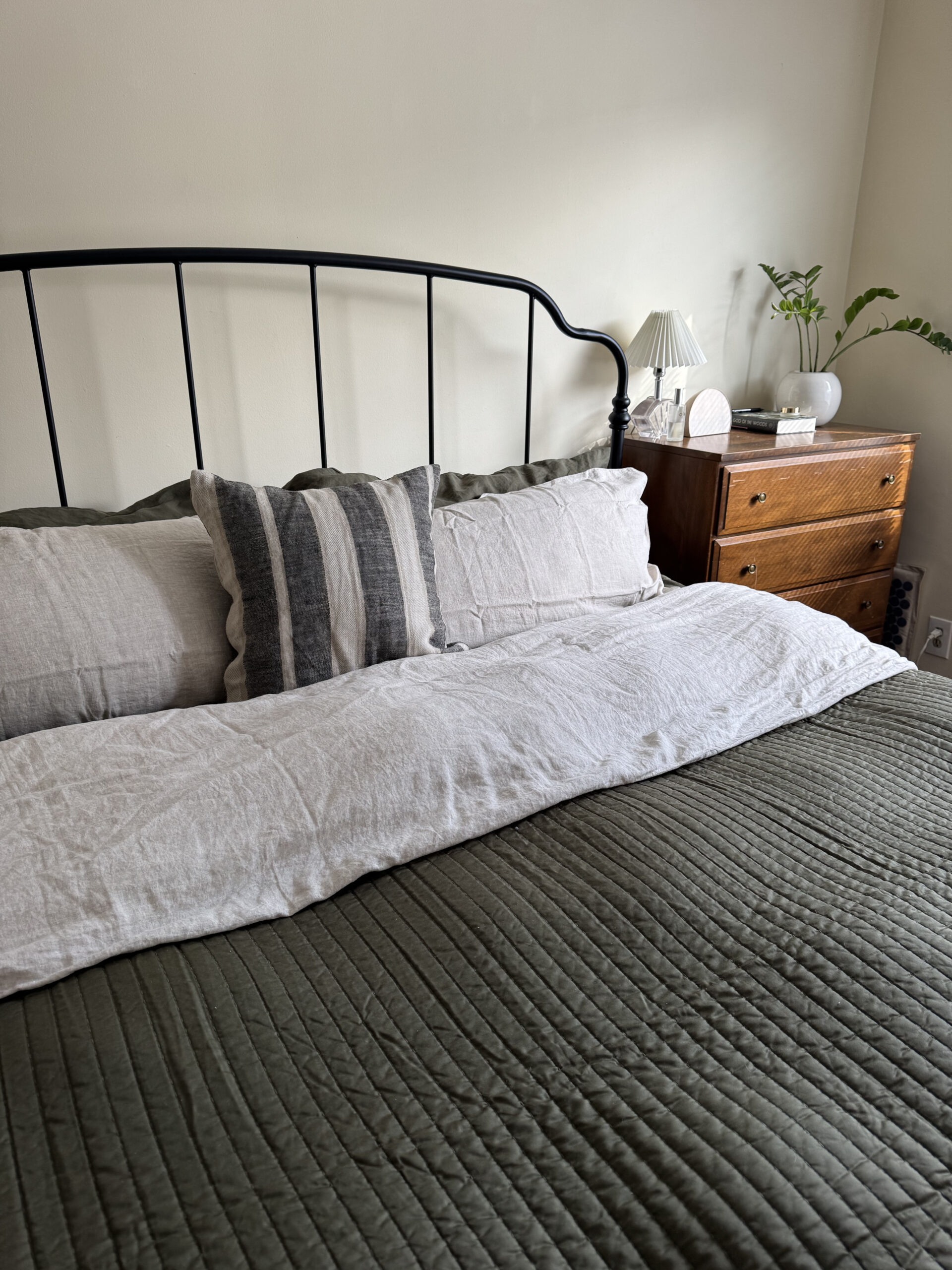 A neatly made bed with green quilt, light-colored pillows, and a black metal headboard. A wooden nightstand holds a small lamp and decorative items.