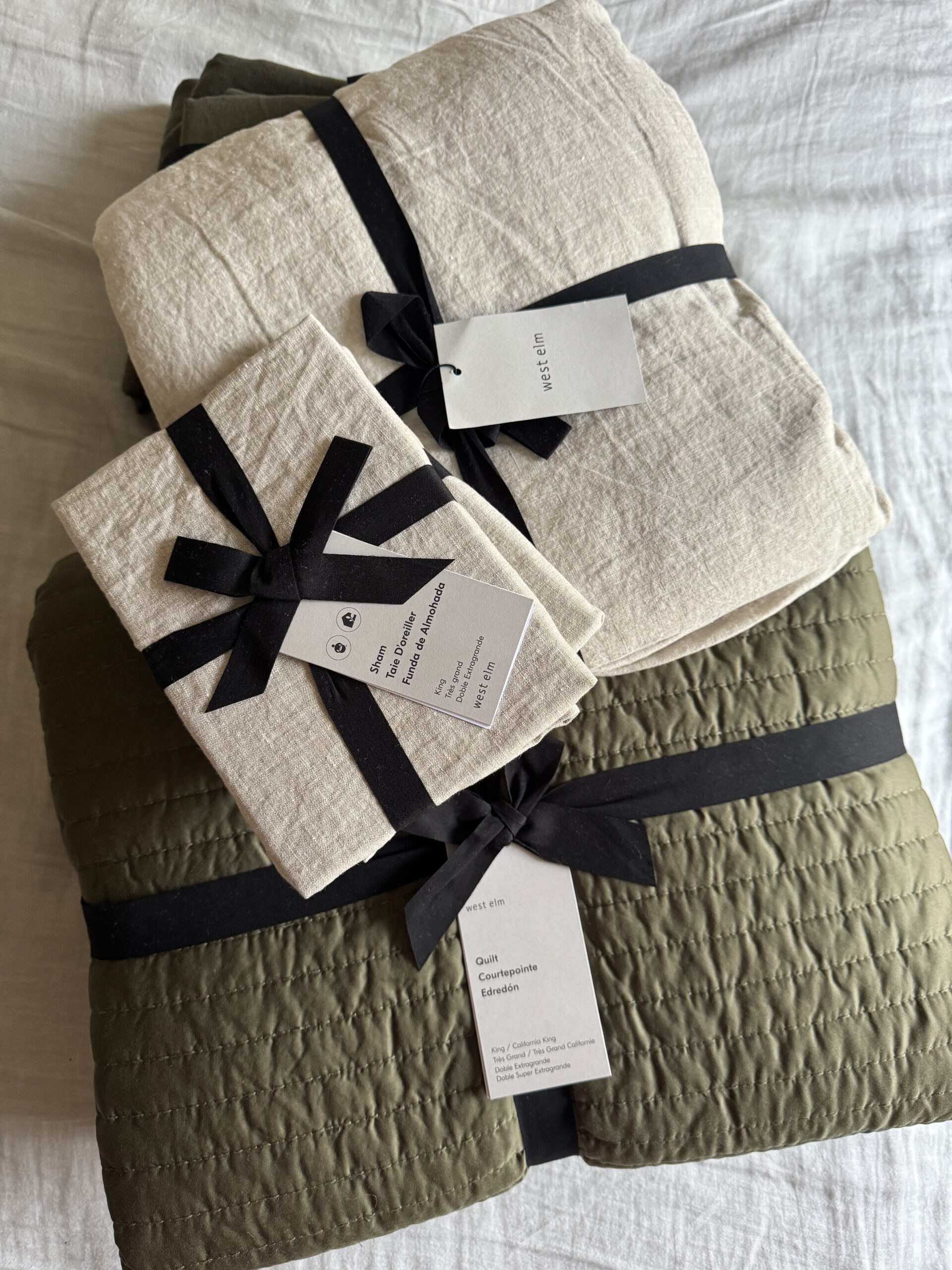 A stack of folded neutral-colored bedding with black ribbon and tags, placed on a white fabric surface.