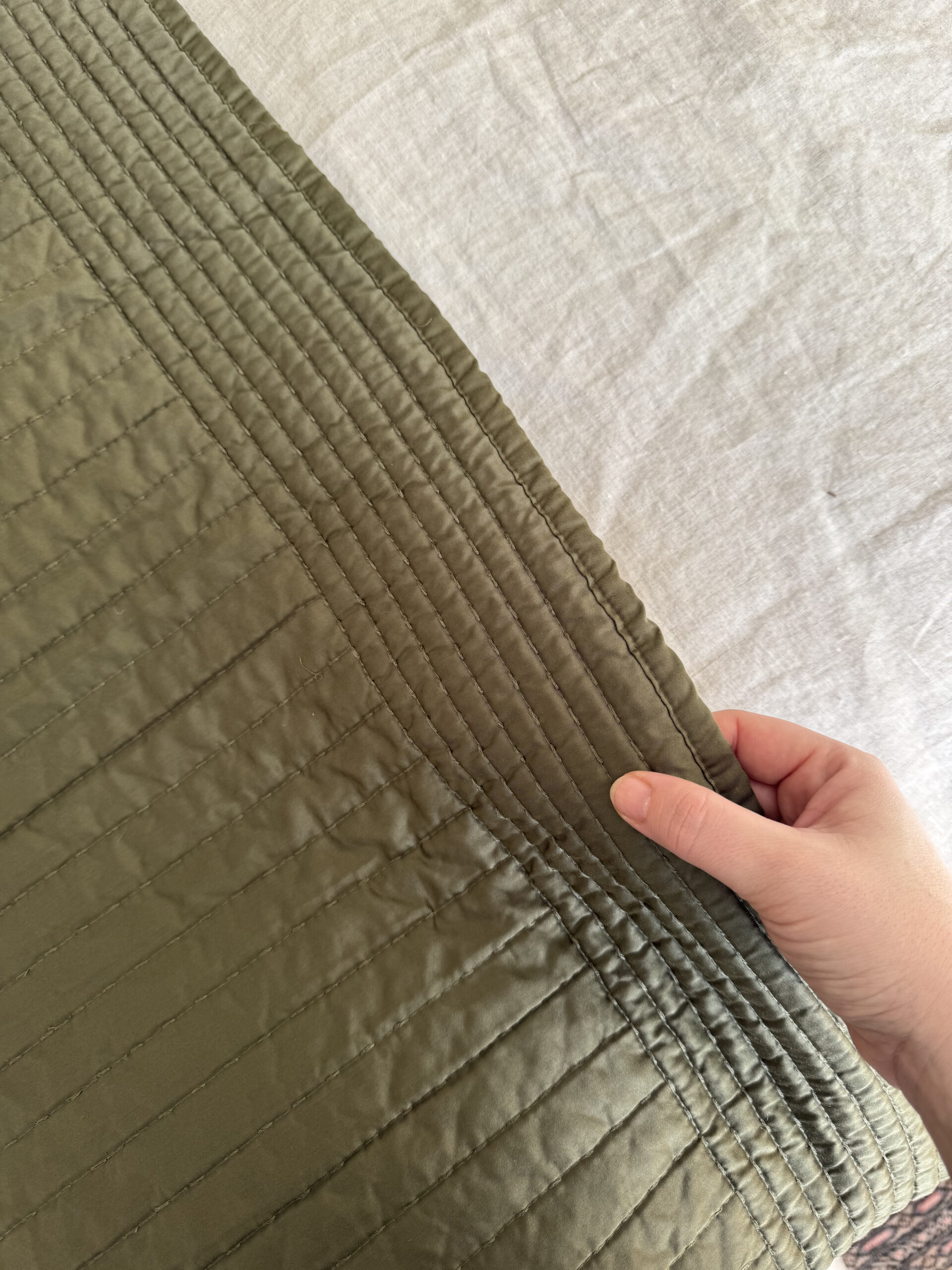 A hand holds the corner of a quilted fabric with parallel stitching. The fabric is a smooth, olive green material against a light-colored textured background.