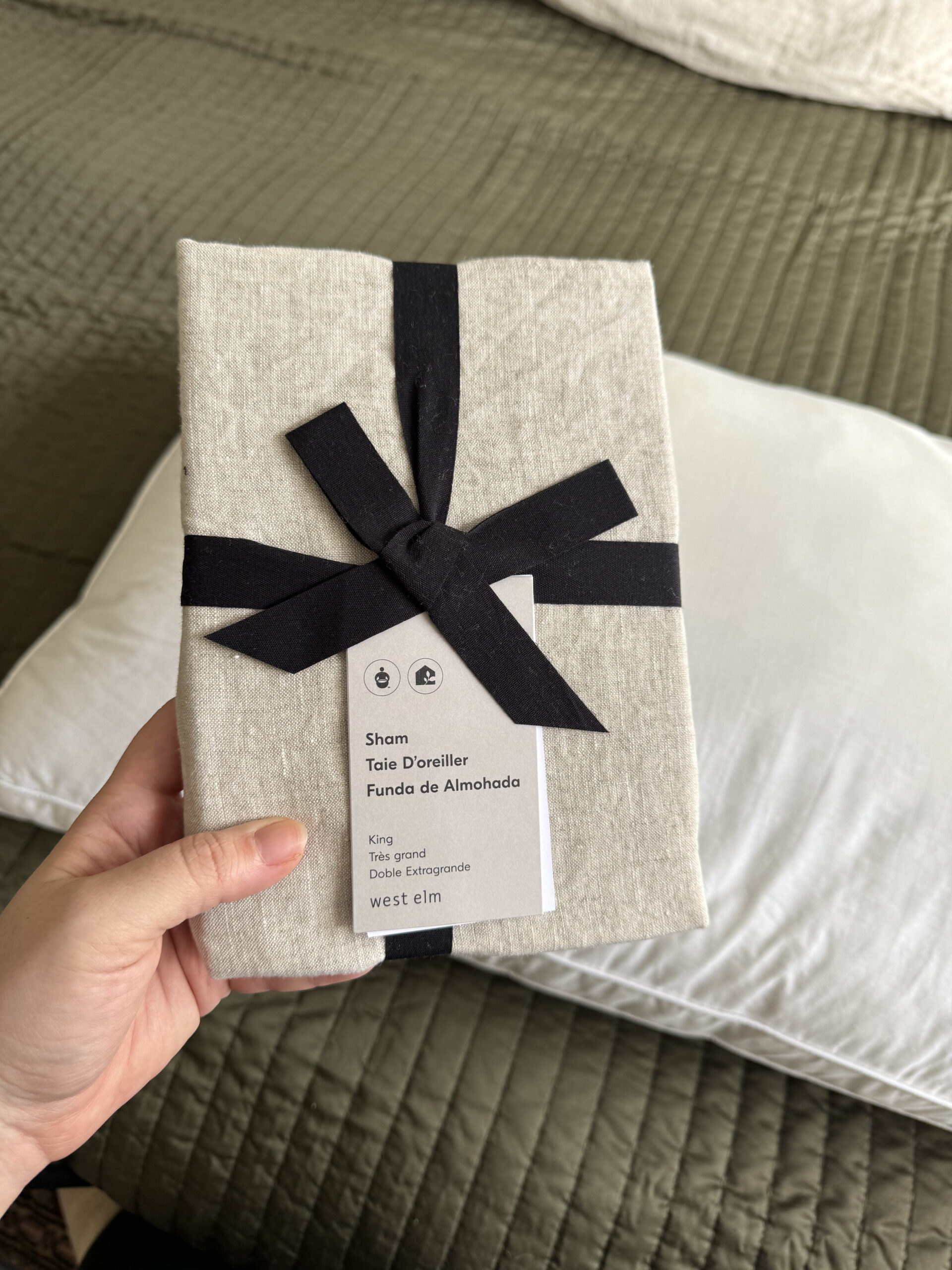 A hand holding a neatly wrapped pillow sham package with a black ribbon. The package label indicates it's a king size pillow sham from West Elm. A pillow and green bedding are in the background.