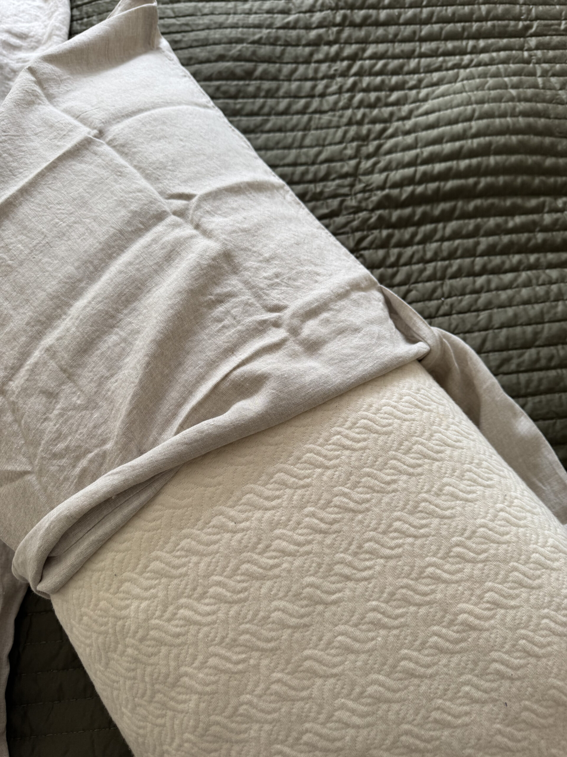 Close-up of a white textured pillow partially covered by a white sheet, on top of a dark green quilted bedspread.