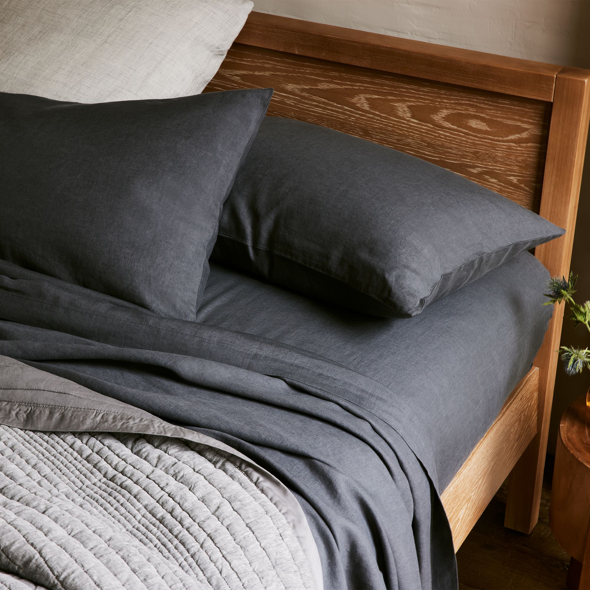 A neatly made bed with dark gray sheets and pillows on a wooden frame. A light gray quilted blanket is folded at the foot of the bed.