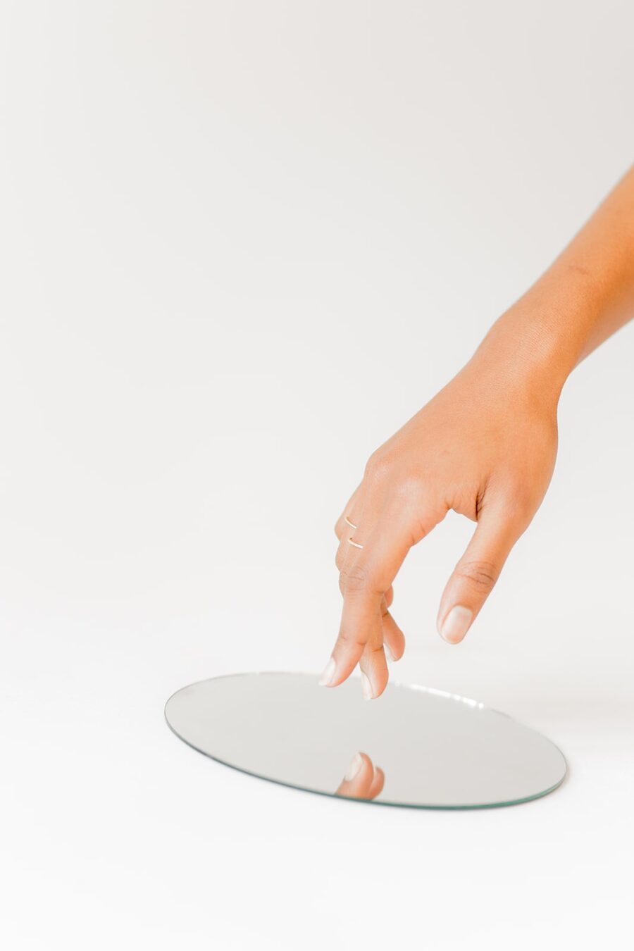 Hand reaching towards a round mirror lying flat on a white surface, with fingers gently touching the edge.