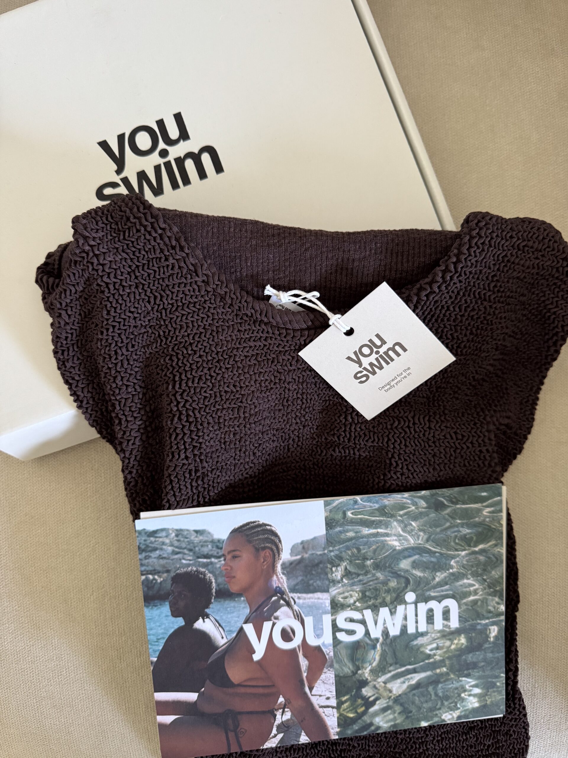 A textured brown swimsuit with a "youswim" tag, a branded box, and a postcard featuring two women in swimwear near water.