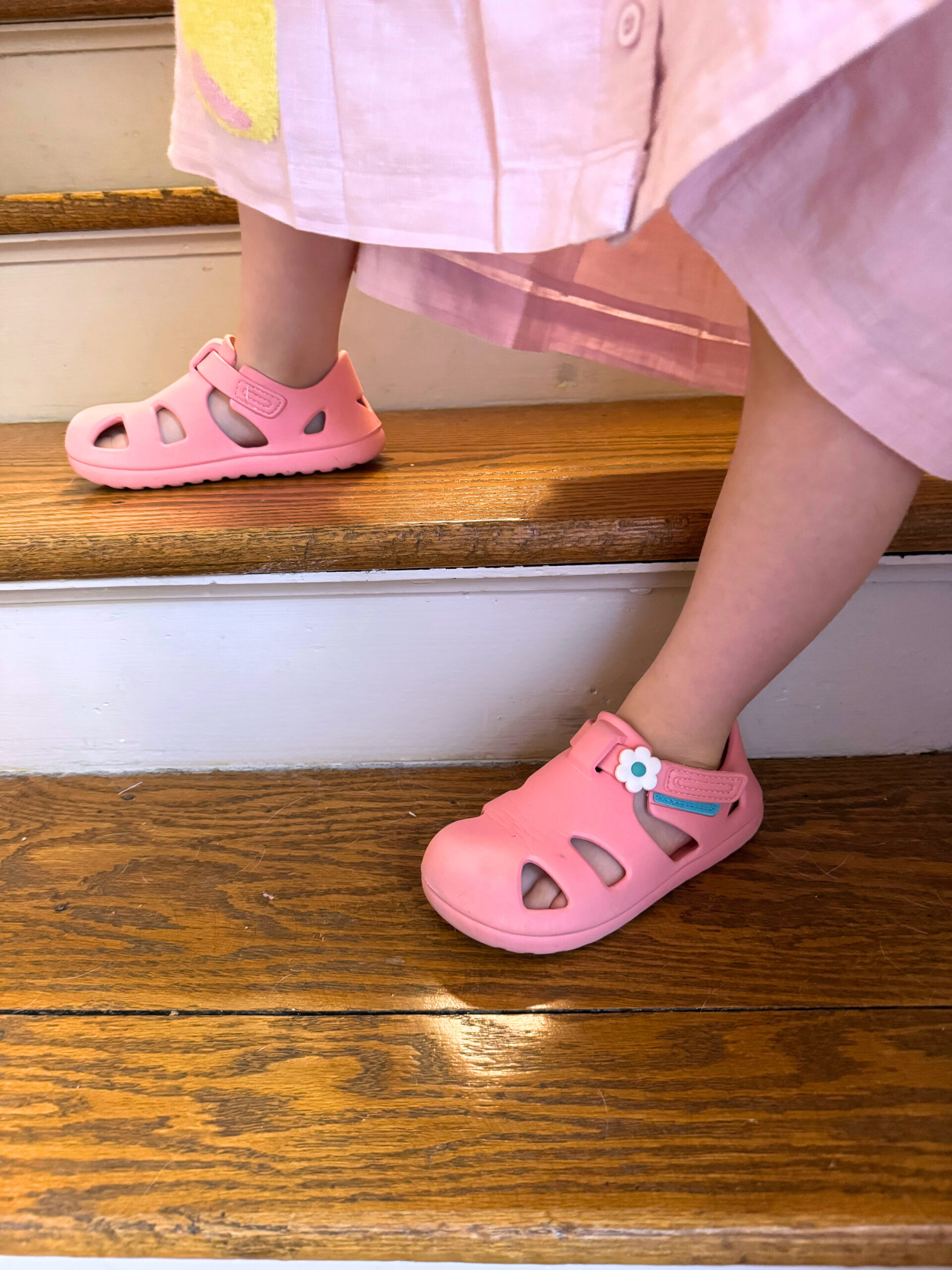 A child wearing a pink dress and pink sandals is stepping up wooden stairs.