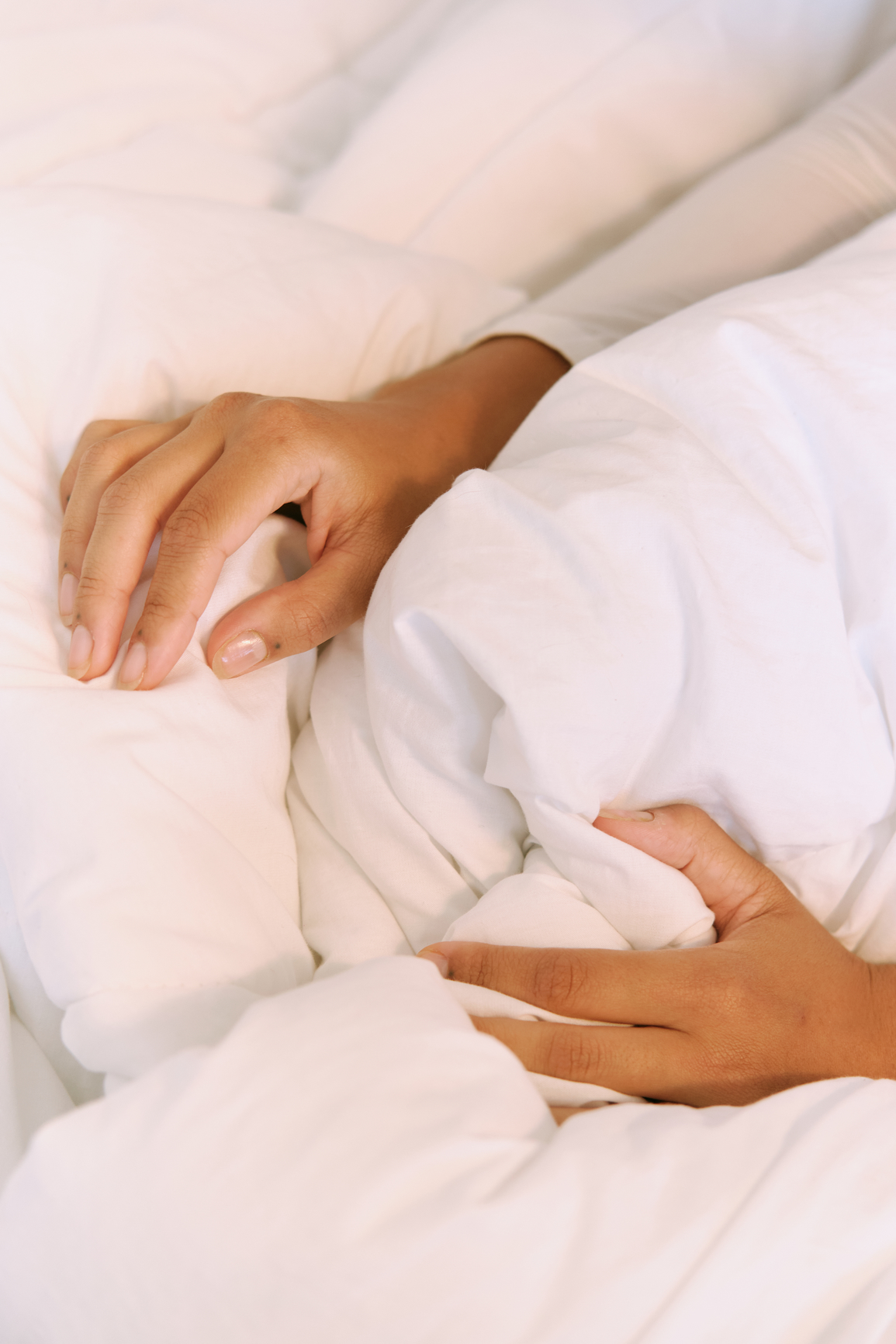 A person’s hands gently hold and gather white bed sheets, creating folds and layers in the fabric.