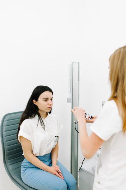 A woman sits in a gray chair, facing another woman standing with medical equipment in a well-lit, white room.