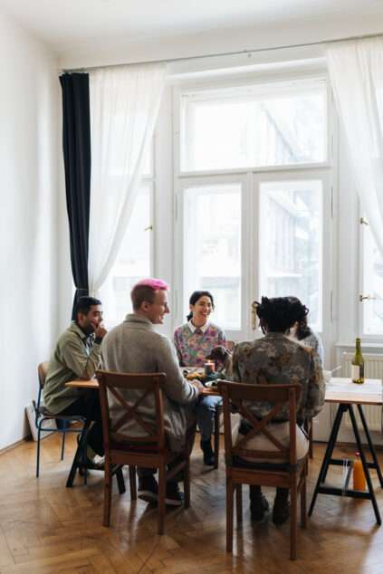 A group of five people sit around a table near a large window in a bright room, engaged in conversation.