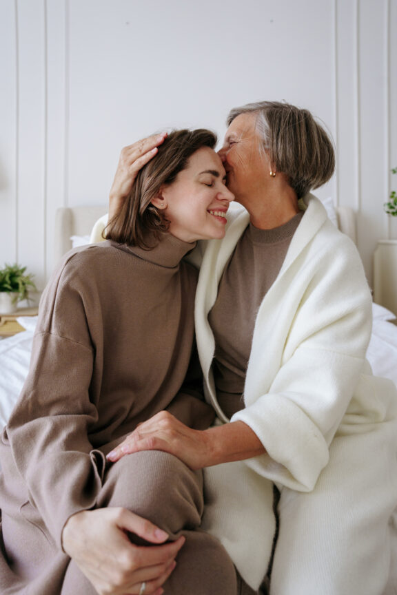 Two women sitting on a bed; one older woman kisses the younger woman's forehead. They both wear cozy, neutral-toned clothing.