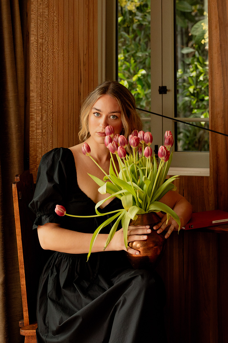 Woman in black dress holding a vase of pink tulips, seated near a window with wooden walls in the background.