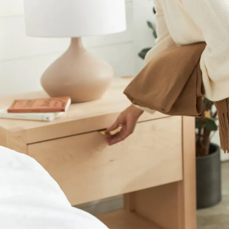 Person holding a brown sweater opens a wooden nightstand drawer beside a table lamp and book.