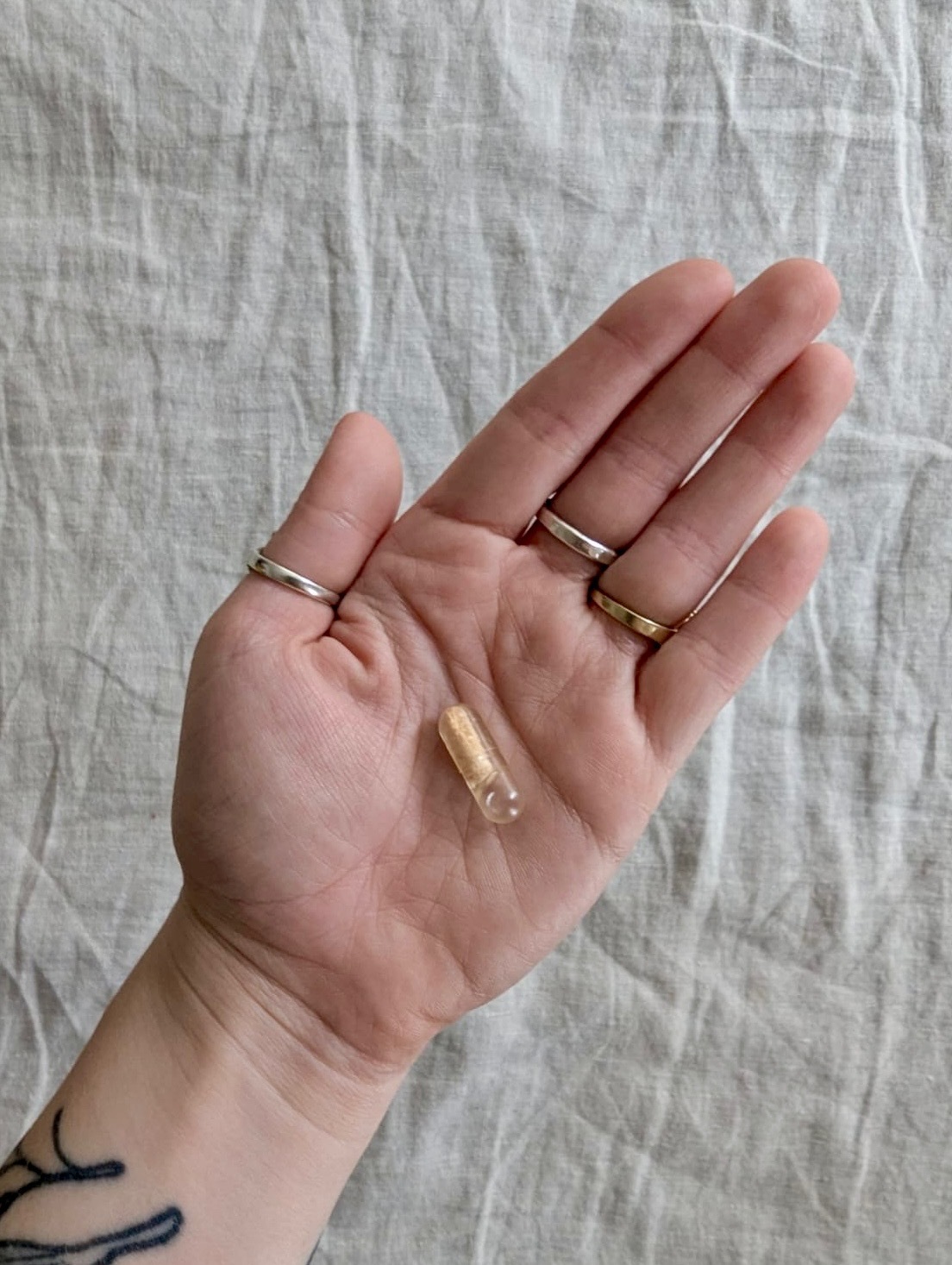 A hand with several rings and a tattoo holds a single clear capsule on the palm against a textured fabric background.