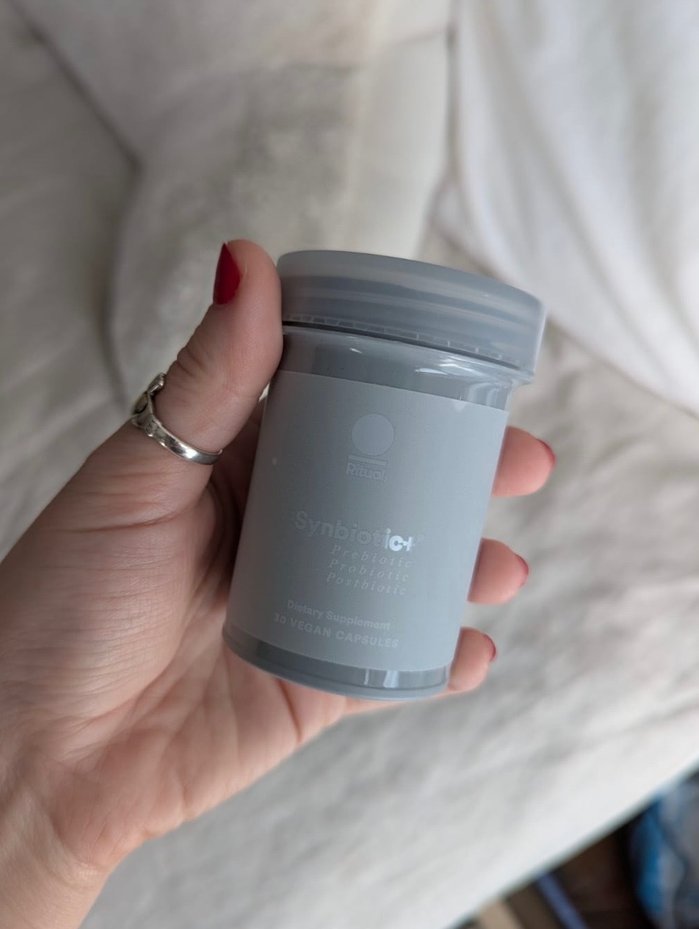 A hand with red nail polish holds a gray bottle labeled "Symbiotic+ Probiotic + Prebiotic + Postbiotic" containing 30 vegan capsules.