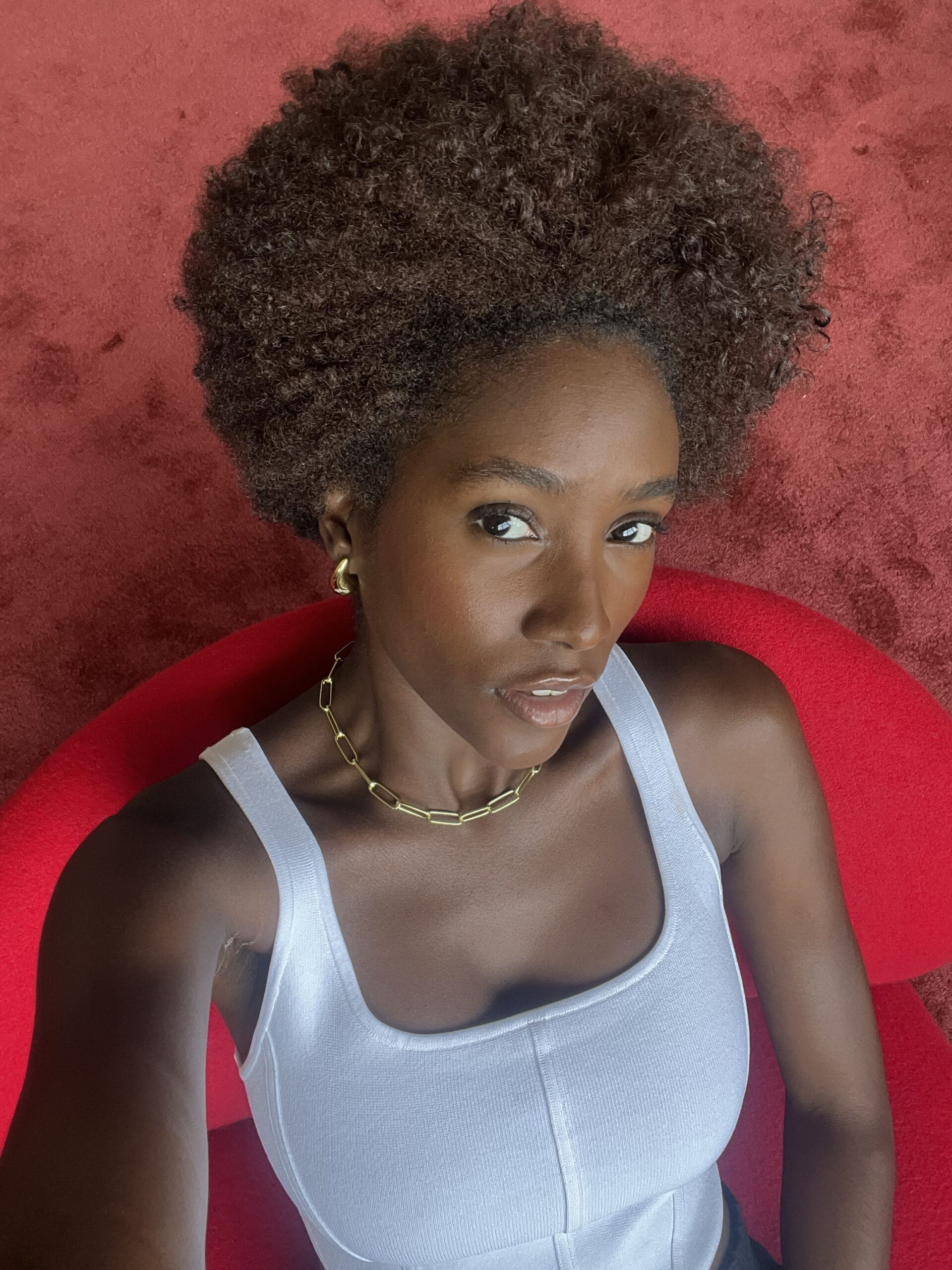 A person with curly hair sits on a red chair, wearing a white tank top and a gold chain necklace, looking at the camera.