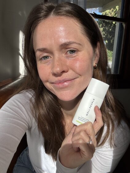 Person smiling and holding a white skincare product bottle indoors near a window.