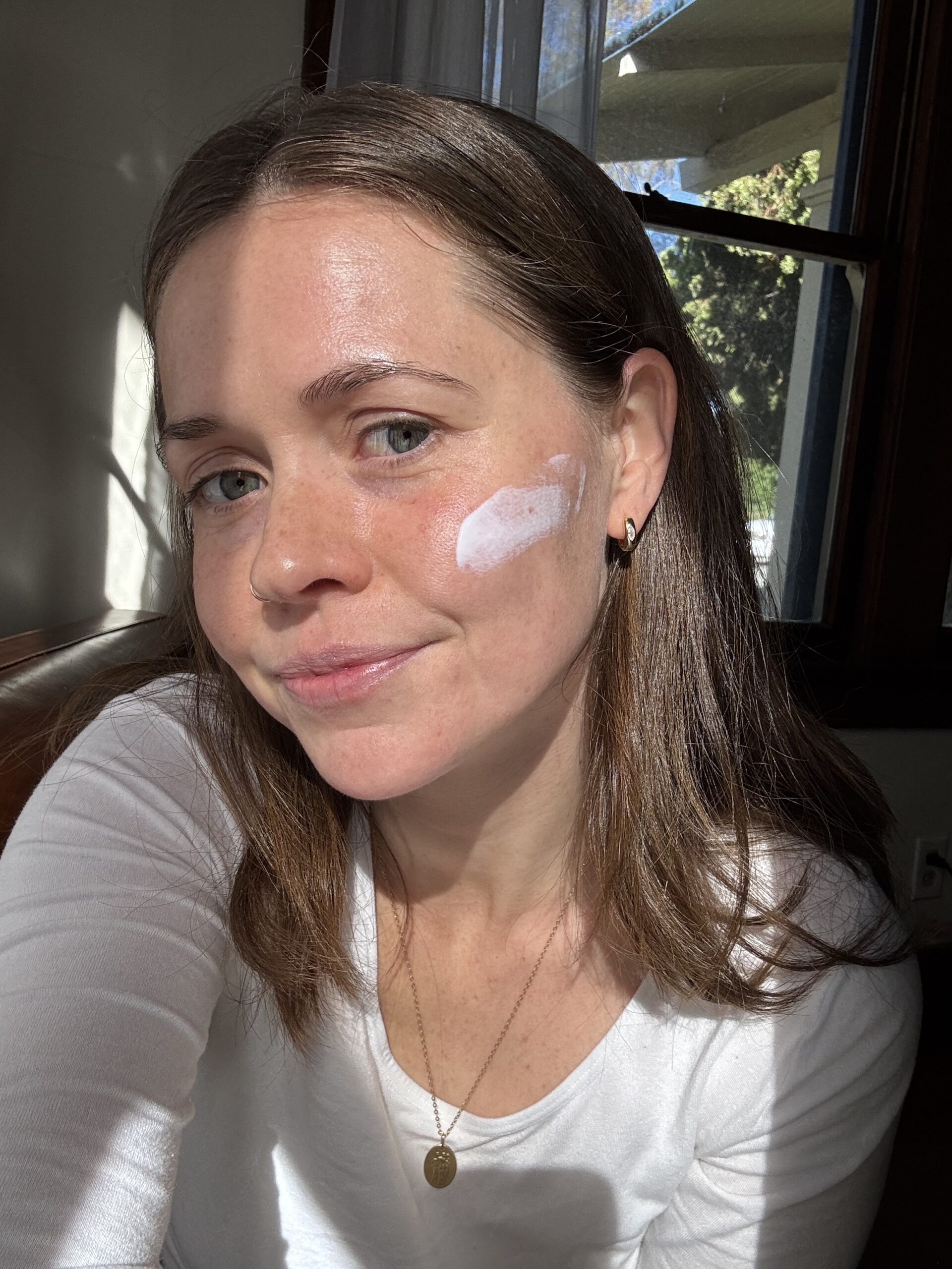 A woman wearing a white shirt has a small amount of cream applied on her cheek. She is sitting indoors by a window with sunlight shining on her.