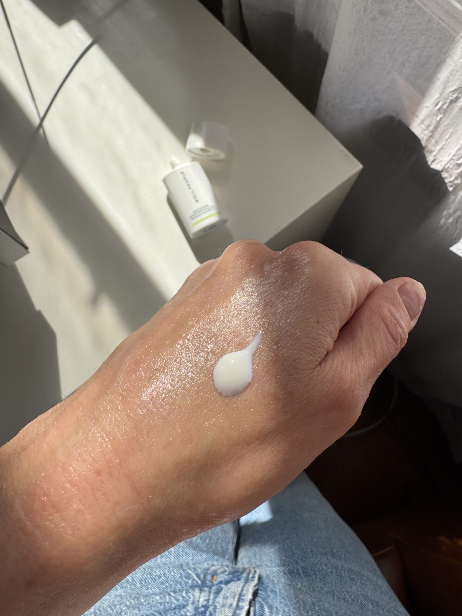 A dollop of white lotion on the back of a hand, with a tube in the background on a white surface.
