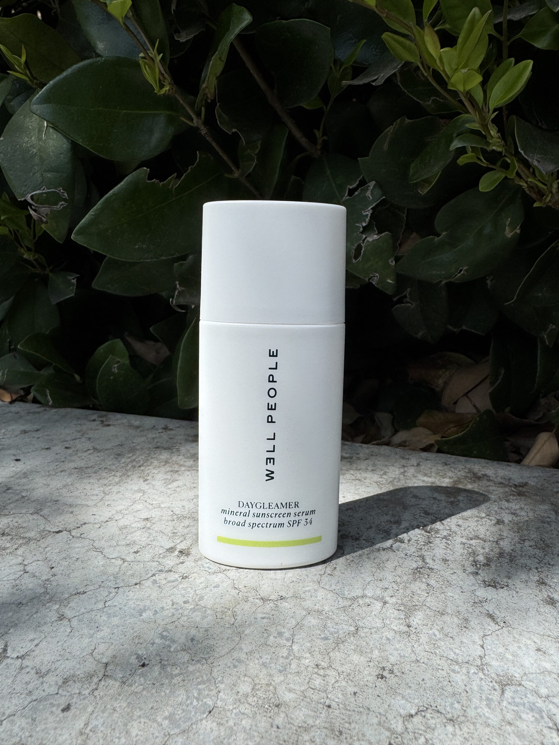 A white bottle of Well People Day Gleamer Broad Spectrum SPF 30 sunscreen stands on a stone surface, surrounded by green foliage.