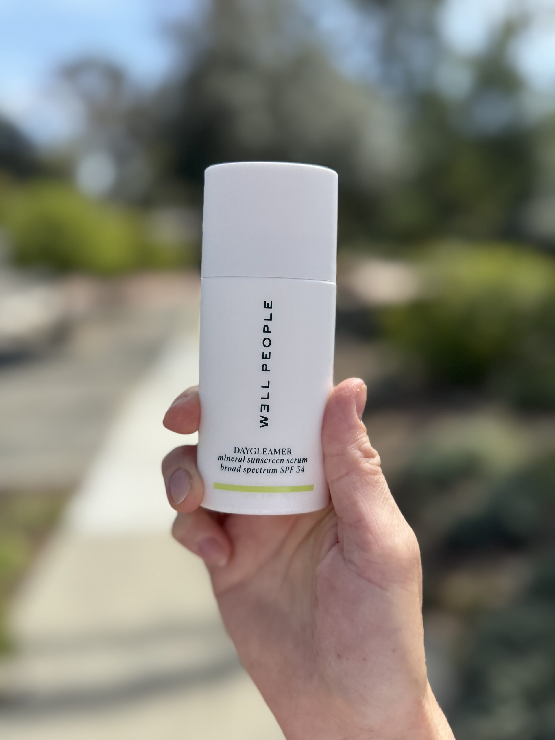 A hand holding a white container labeled "Well People Daydreamer Mineral Sunscreen, broad spectrum SPF 30" with a blurred outdoor background.