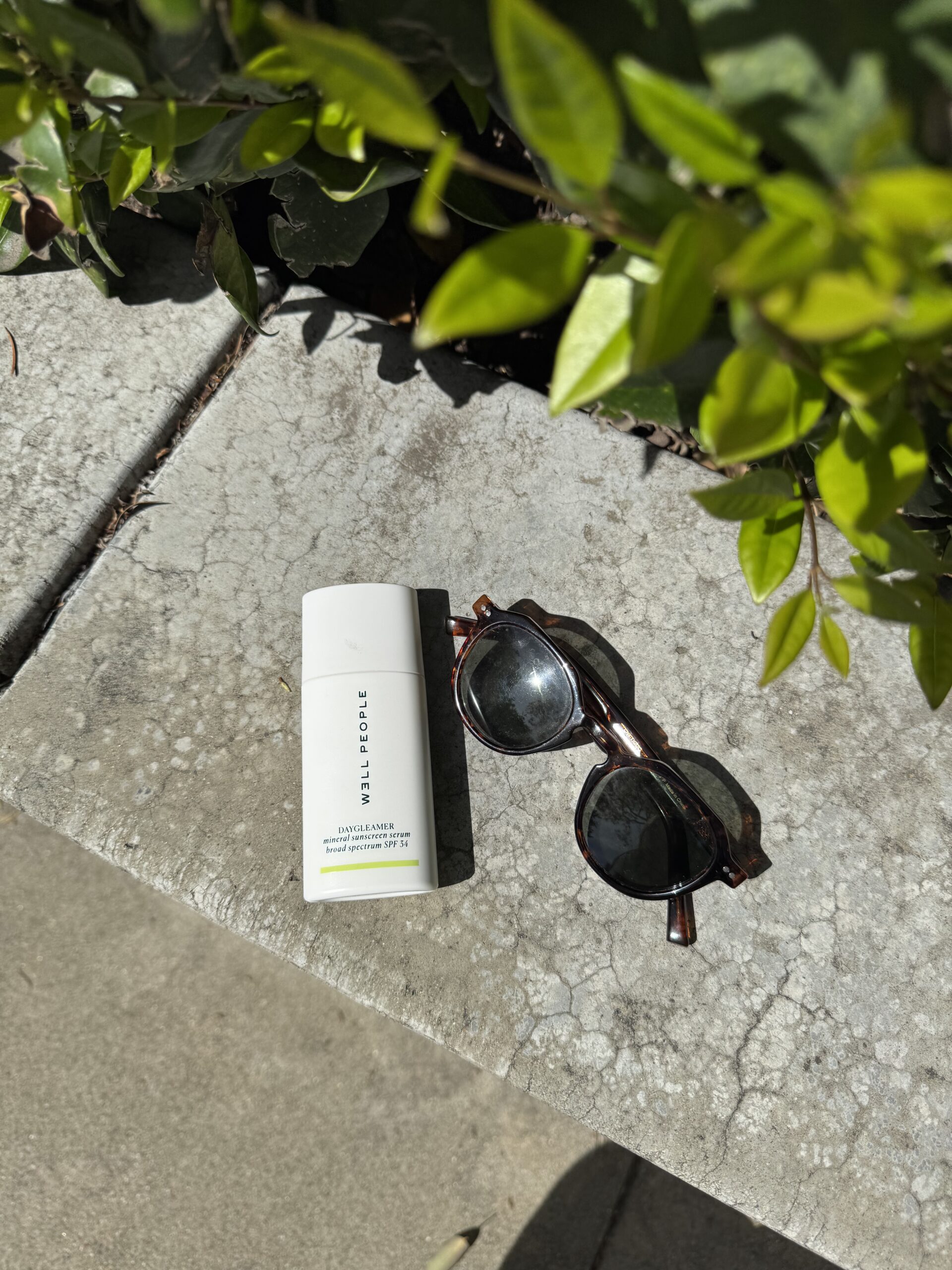 A tube of sunscreen and a pair of round sunglasses on a concrete surface beside green leaves in bright sunlight.