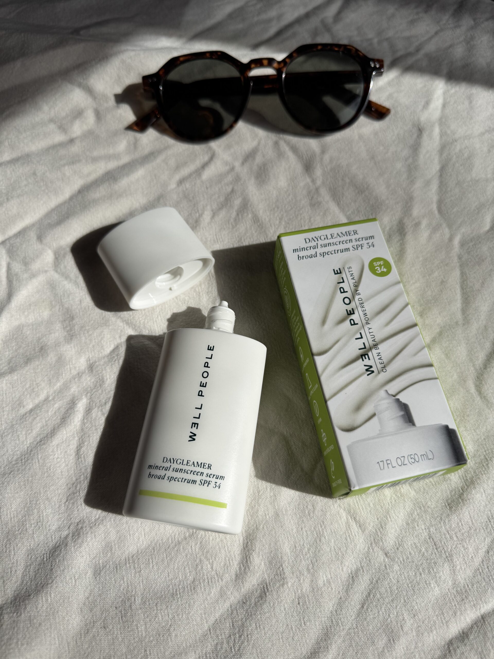Sunglasses next to an open bottle and box of Well People Daydreamer mineral sunscreen SPF 30 on a cloth surface.