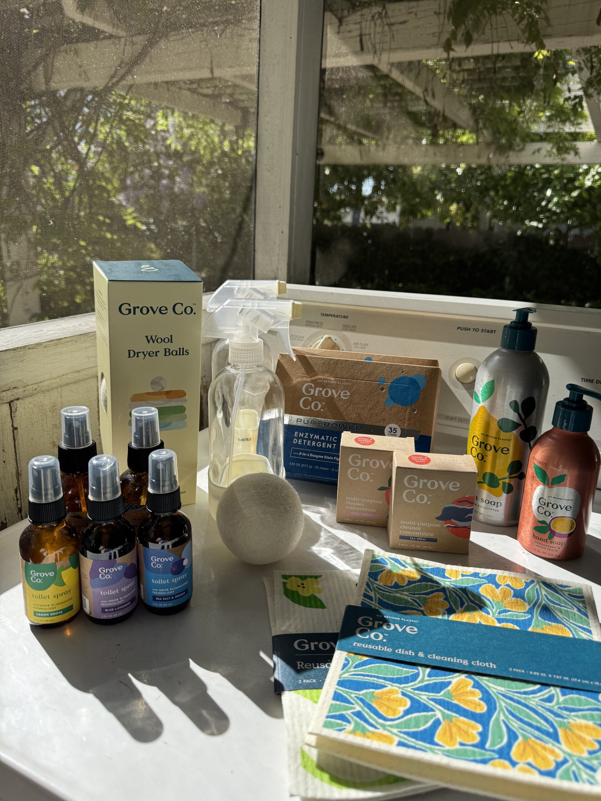 A selection of Grove Co. eco-friendly household products, including soaps, sprays, cleaning concentrates, dryer balls, and dish cloths, displayed on a sunlit table.