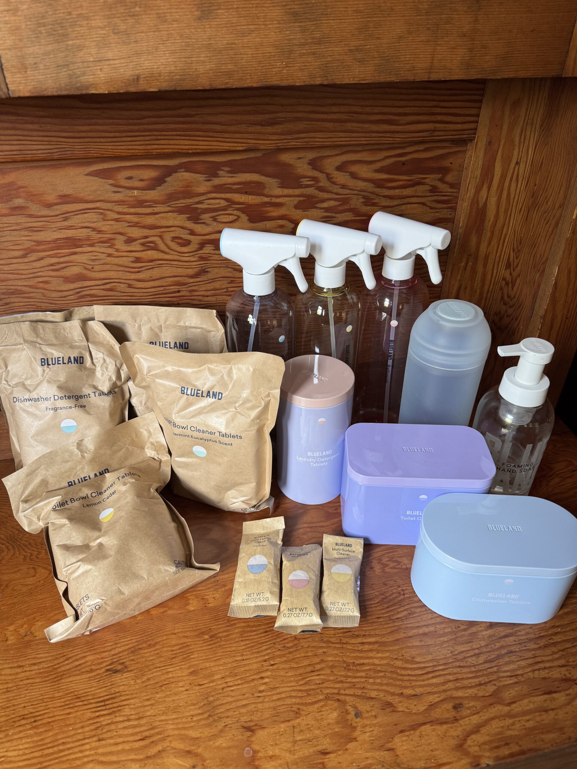 A collection of Blueland cleaning products, including spray bottles, tablets in paper packets, foaming soap dispenser, and pastel-colored storage tins, arranged on a wooden surface.