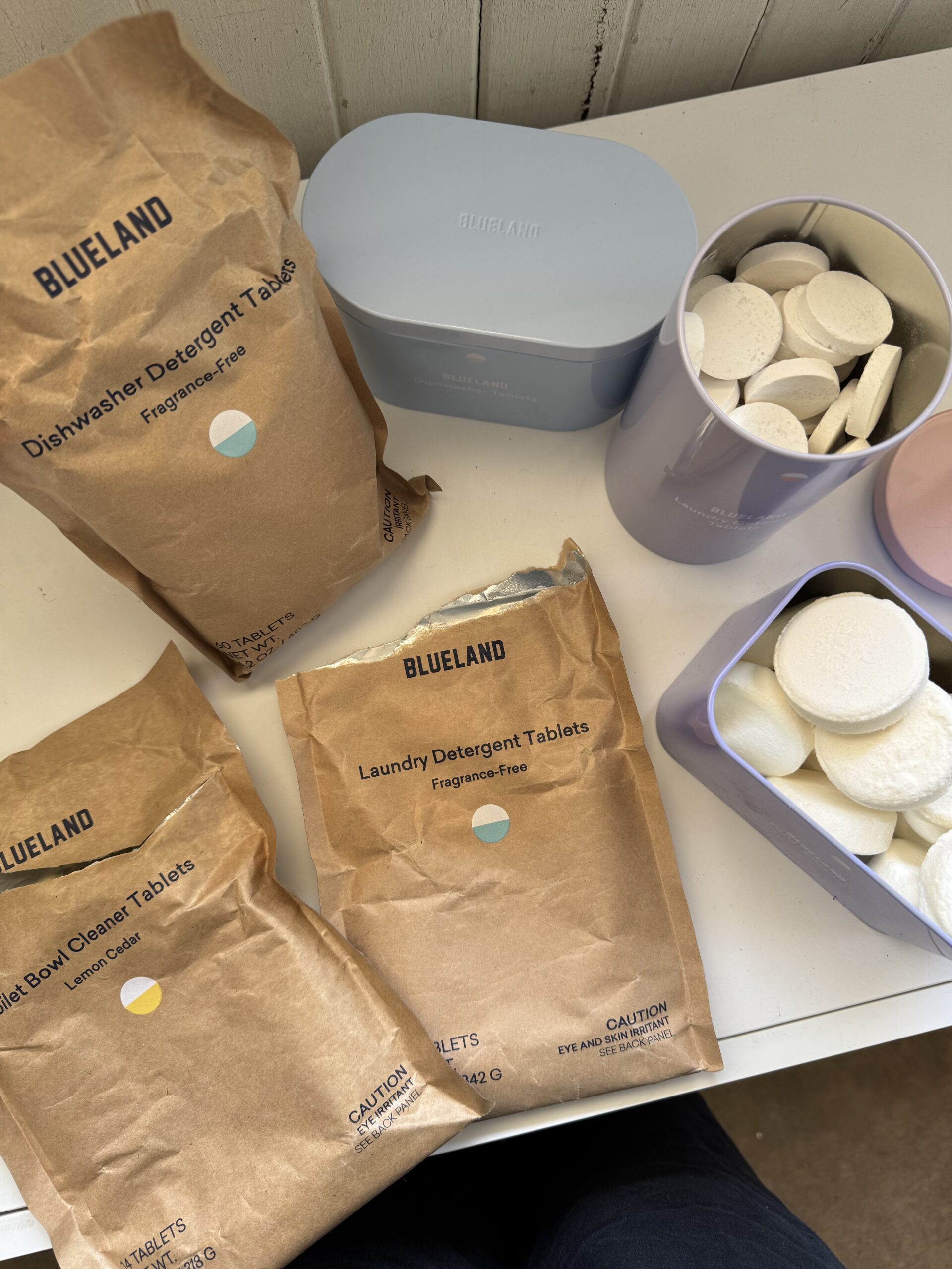 Three brown Blueland detergent tablet bags and two containers with white round tablets are displayed on a white surface.