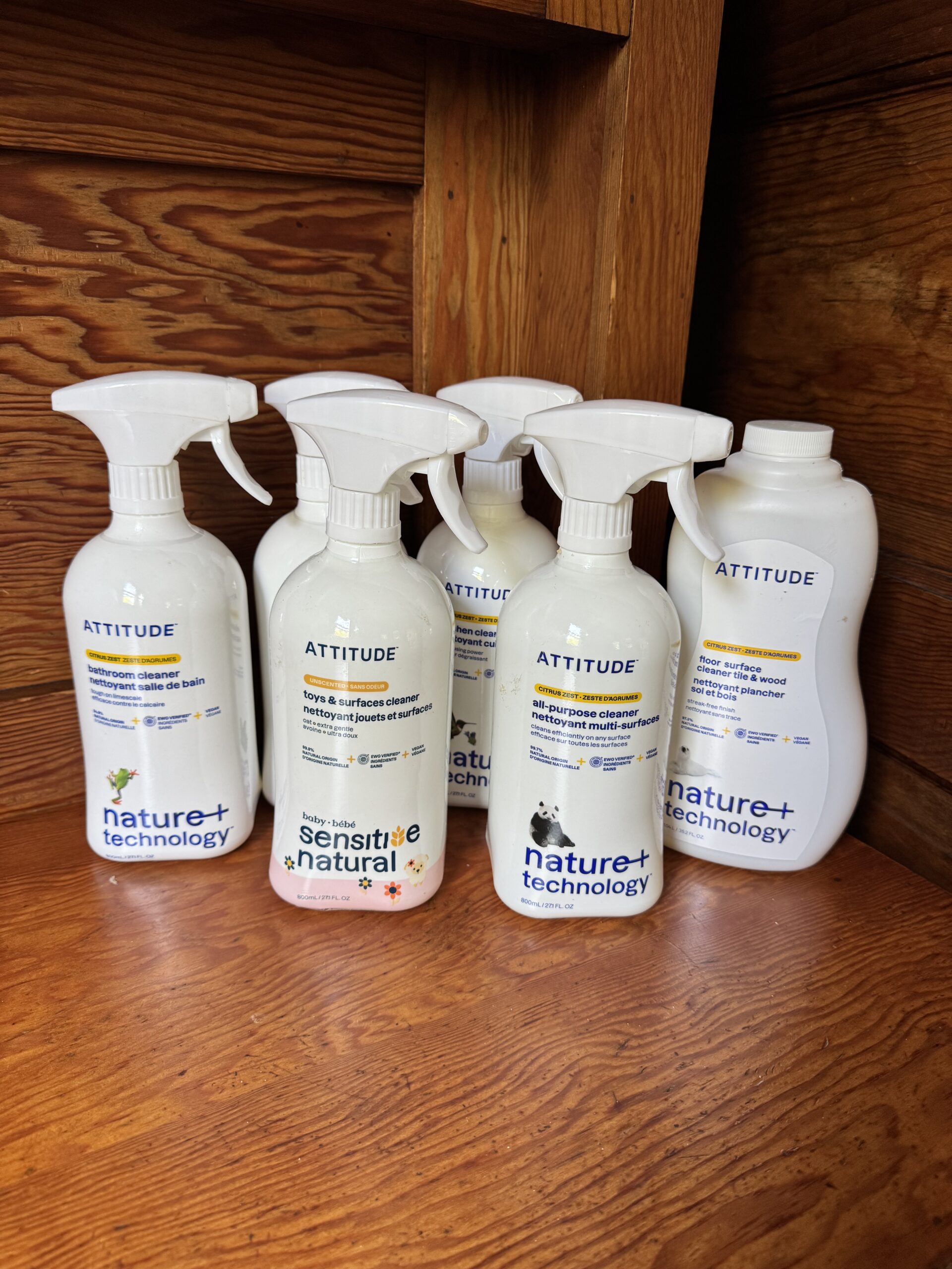 Five bottles of Attitude Nature+ Technology cleaning products are arranged on a wooden shelf, including sensitive and natural formulas in spray and pour bottles.