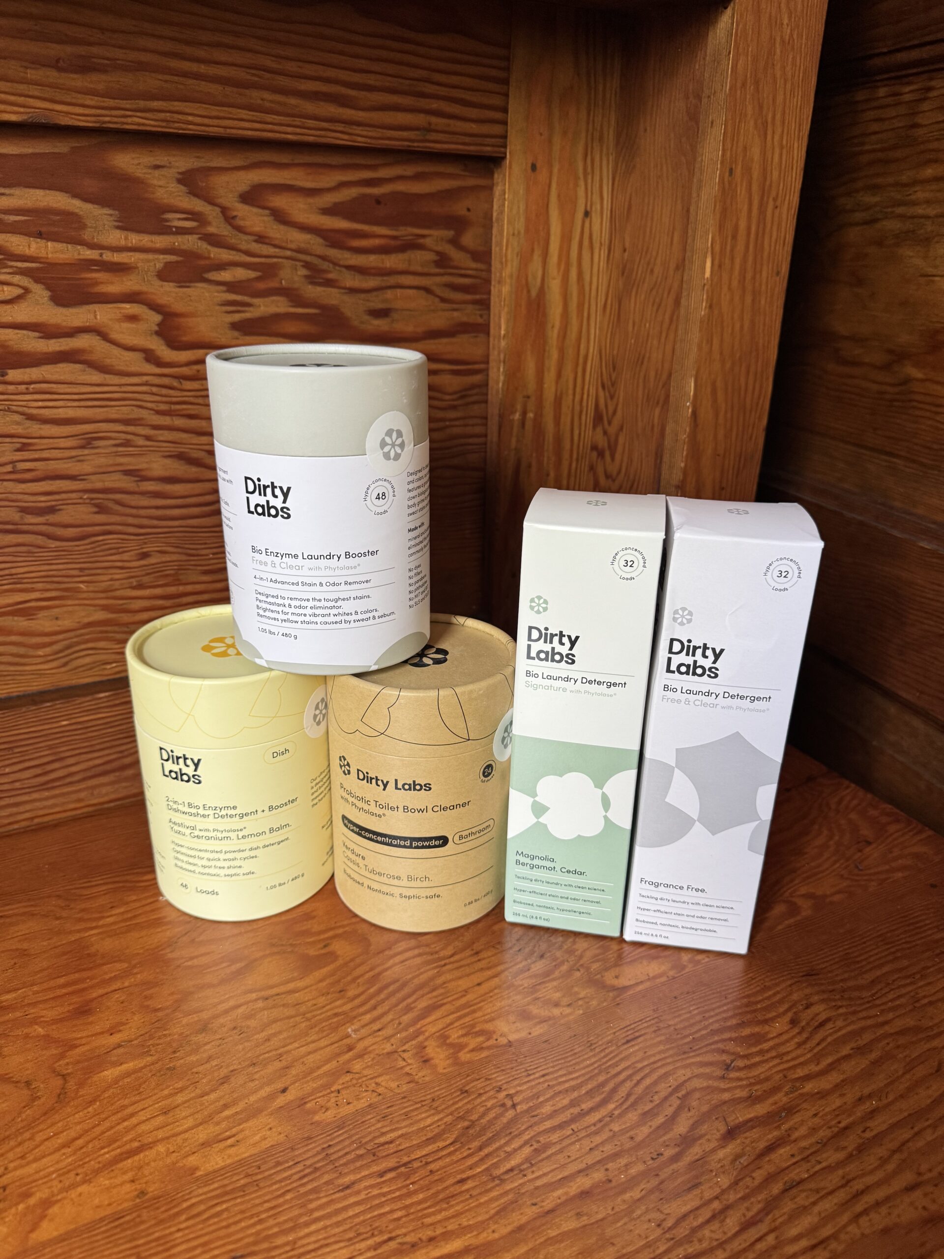 Five Dirty Labs laundry detergent and booster products are arranged on a wooden surface in front of a matching wood-paneled background.