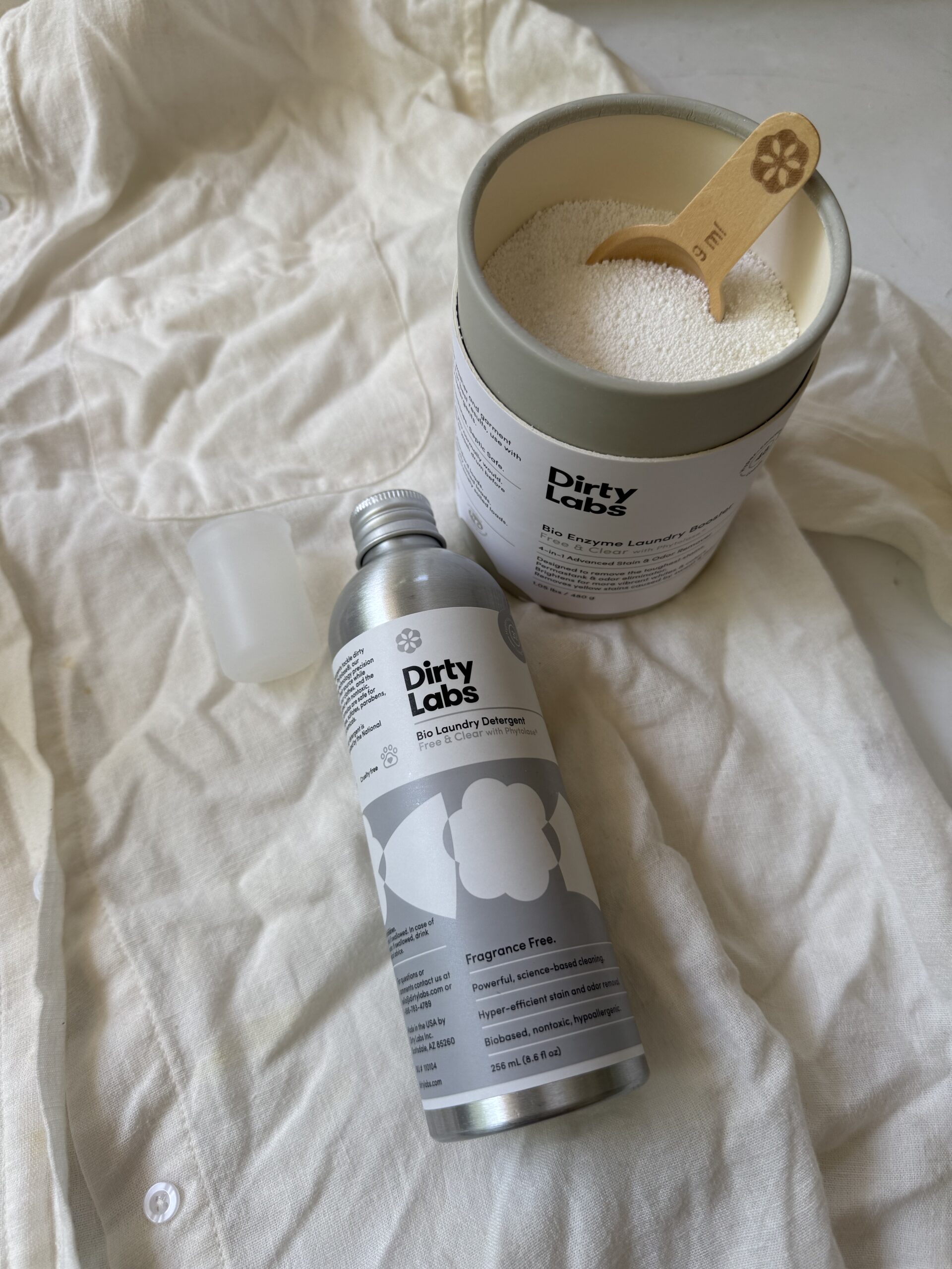 A tin of Dirty Labs laundry powder with a scoop, a bottle of Dirty Labs liquid detergent, and a clear measuring cup on a white fabric background.