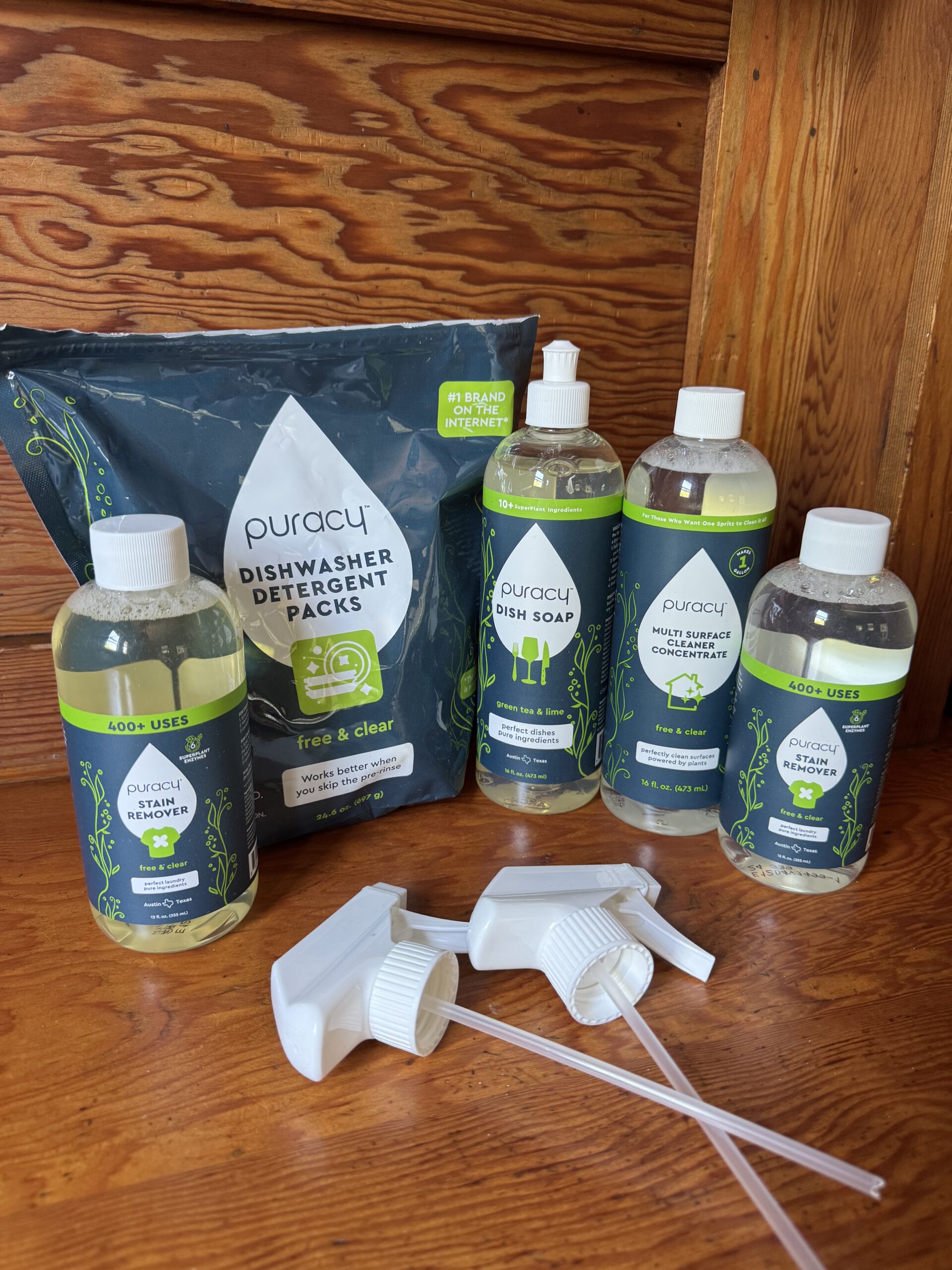 Bottles of Puracy cleaning products and a pack of dishwasher detergent packs are arranged on a wooden surface, with spray nozzles placed in front.