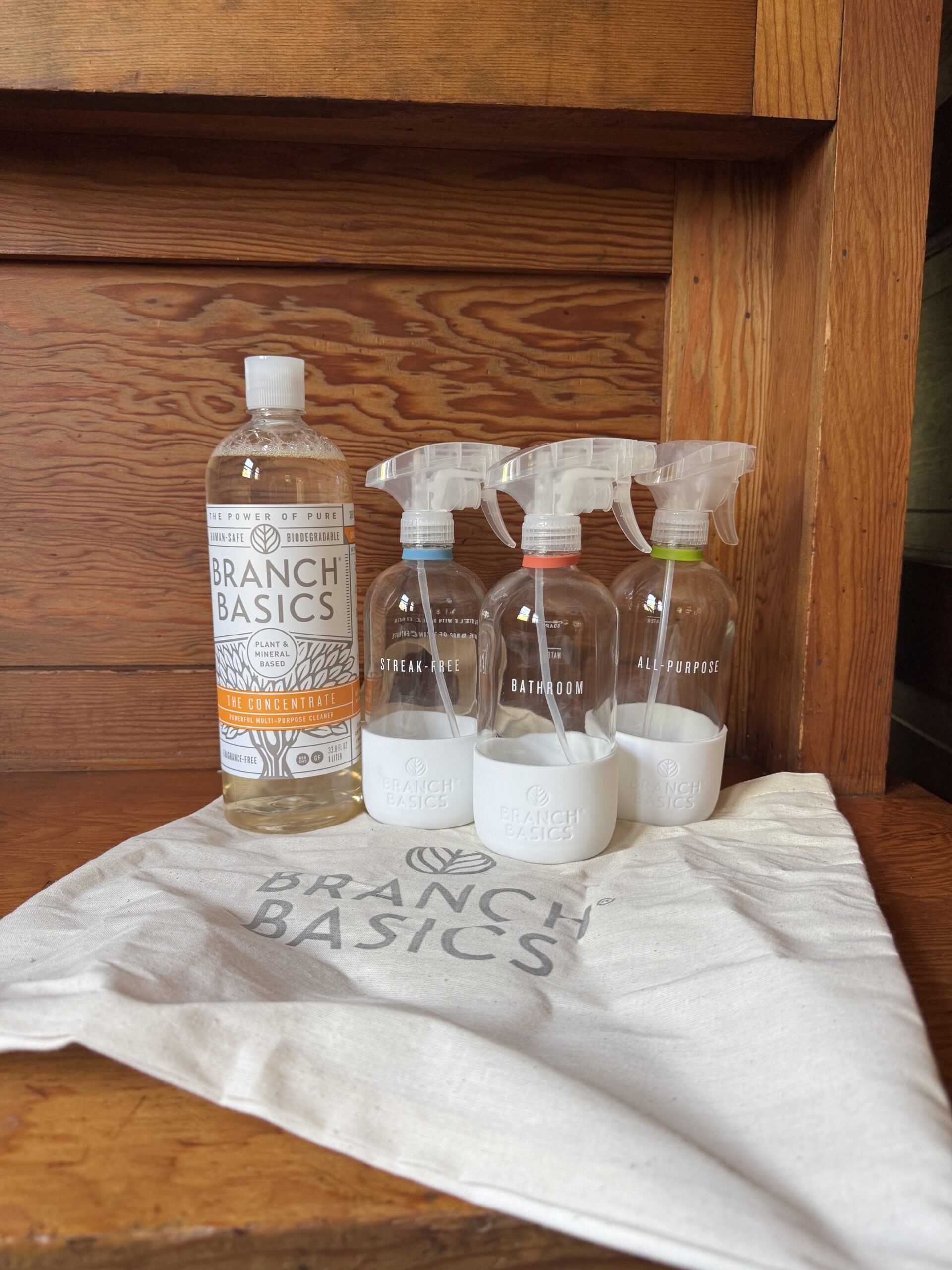 Branch Basics cleaning concentrate bottle and three labeled spray bottles (Streak-Free, Bathroom, All-Purpose) on a branded cloth bag, placed on a wooden surface.