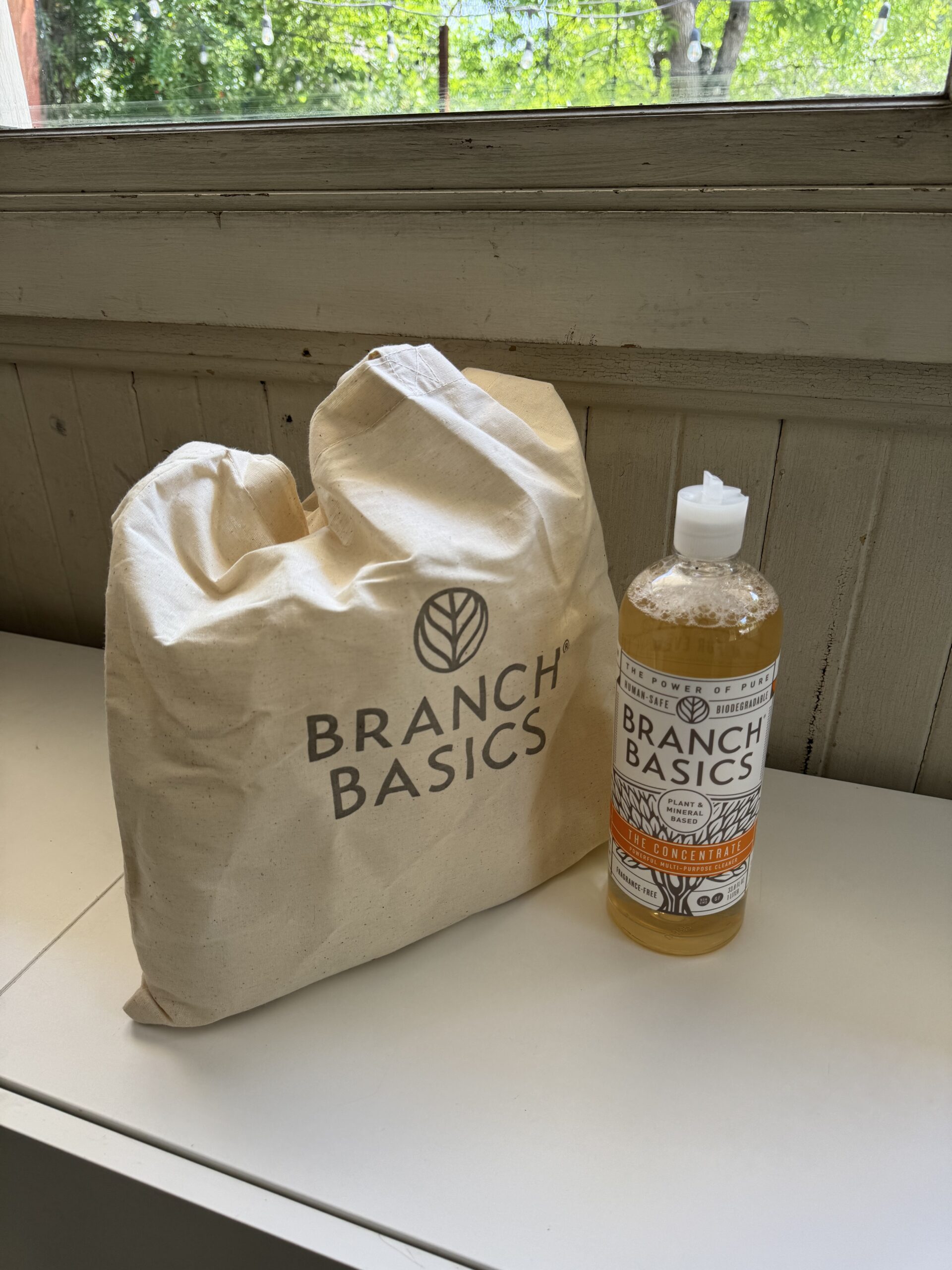 A beige Branch Basics tote bag sits next to a bottle of Branch Basics concentrate on a white surface by a window.