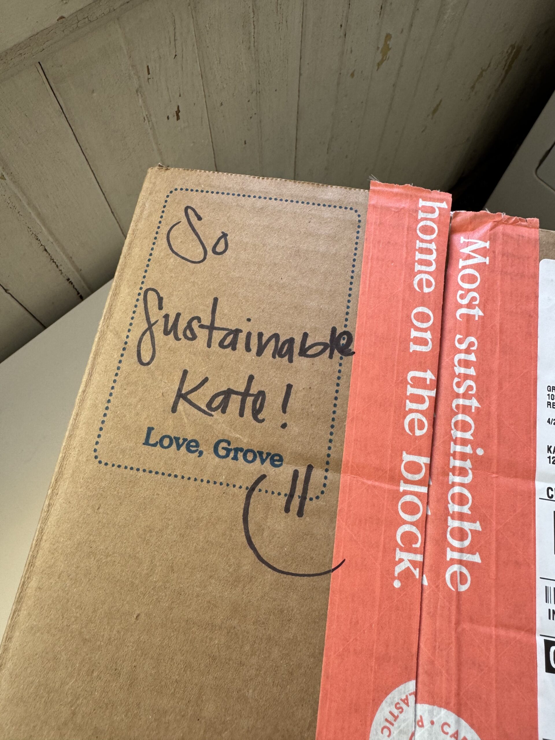 A cardboard shipping box with red tape reading "Most sustainable home on the block." The box has a handwritten note: "So sustainable Kate! Love, Grove" with a smiley face.