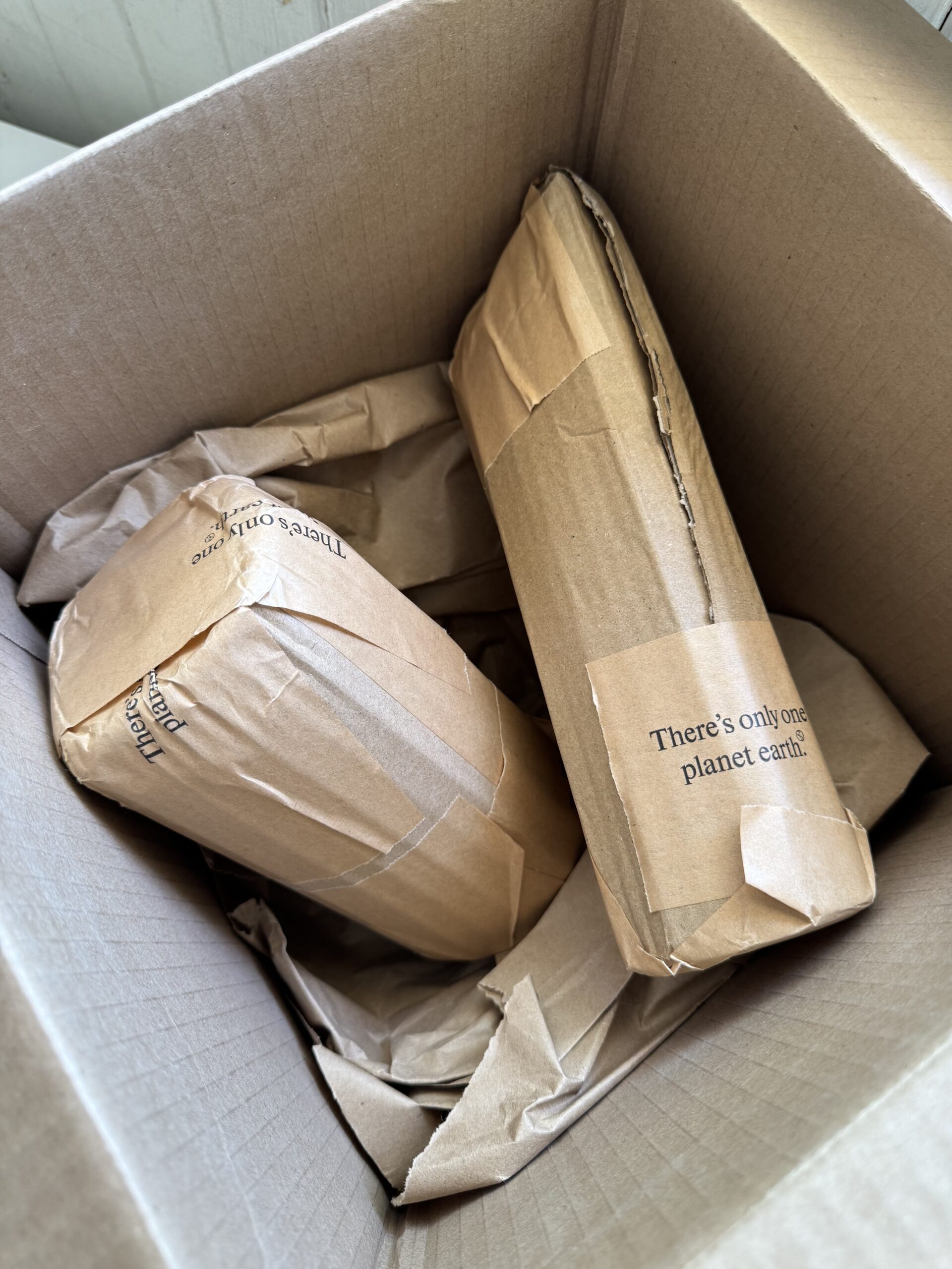 Three items wrapped in brown paper with printed text are packed inside a cardboard box with additional crumpled paper for cushioning.