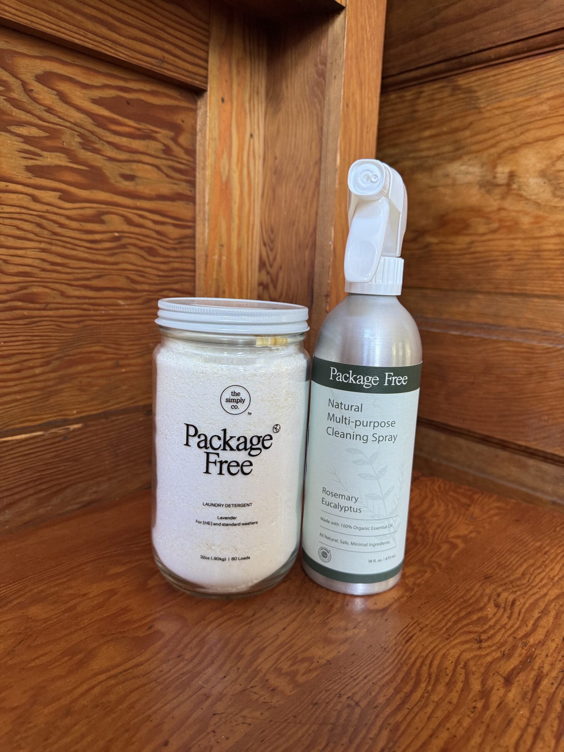 A glass jar of laundry detergent and a spray bottle of multi-purpose cleaning spray, both labeled "Package Free," sit on a wooden corner shelf.