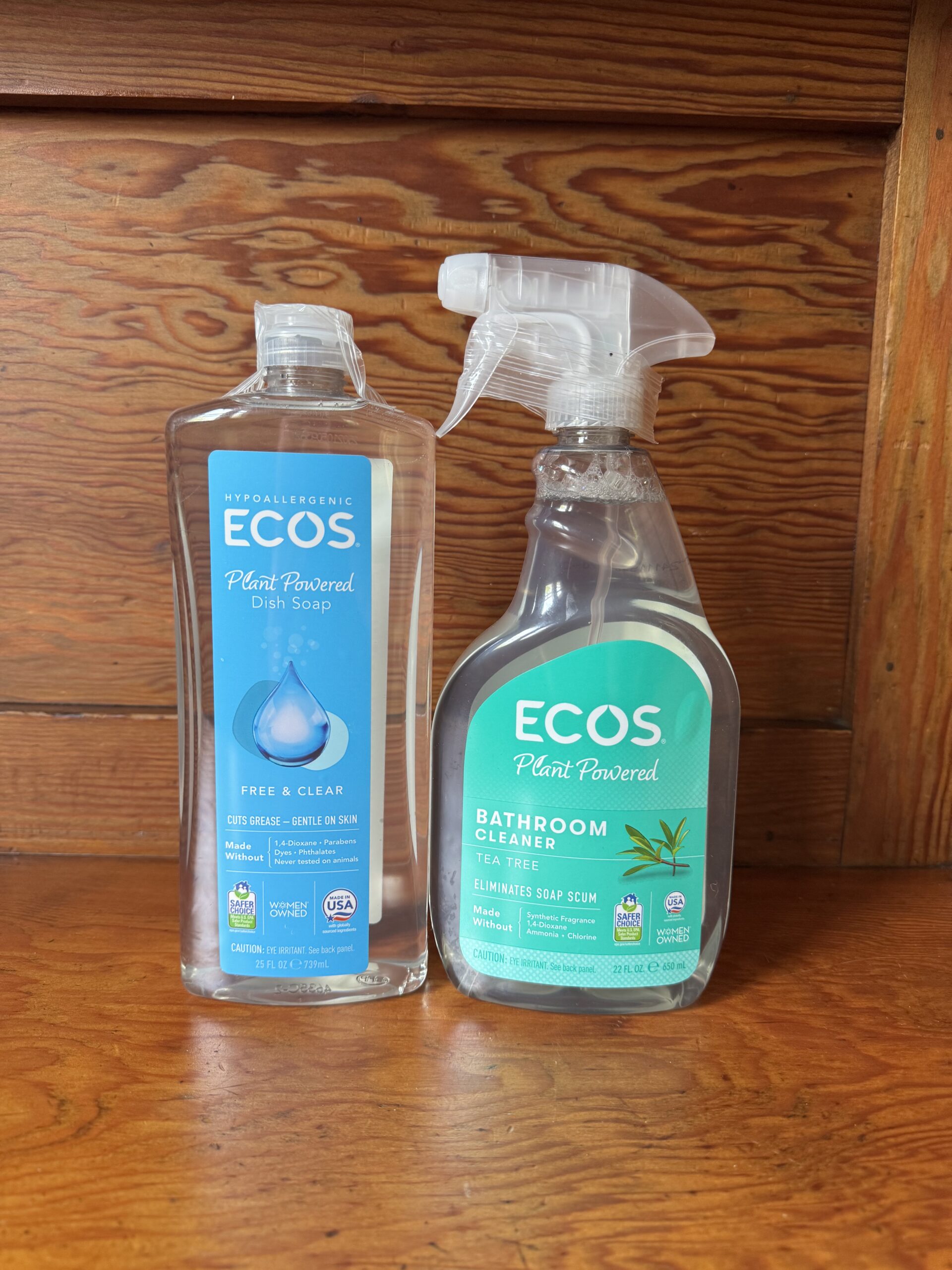 Two bottles of ECOS cleaning products, one dish soap and one bathroom cleaner, are placed side by side on a wooden surface in front of a wooden background.