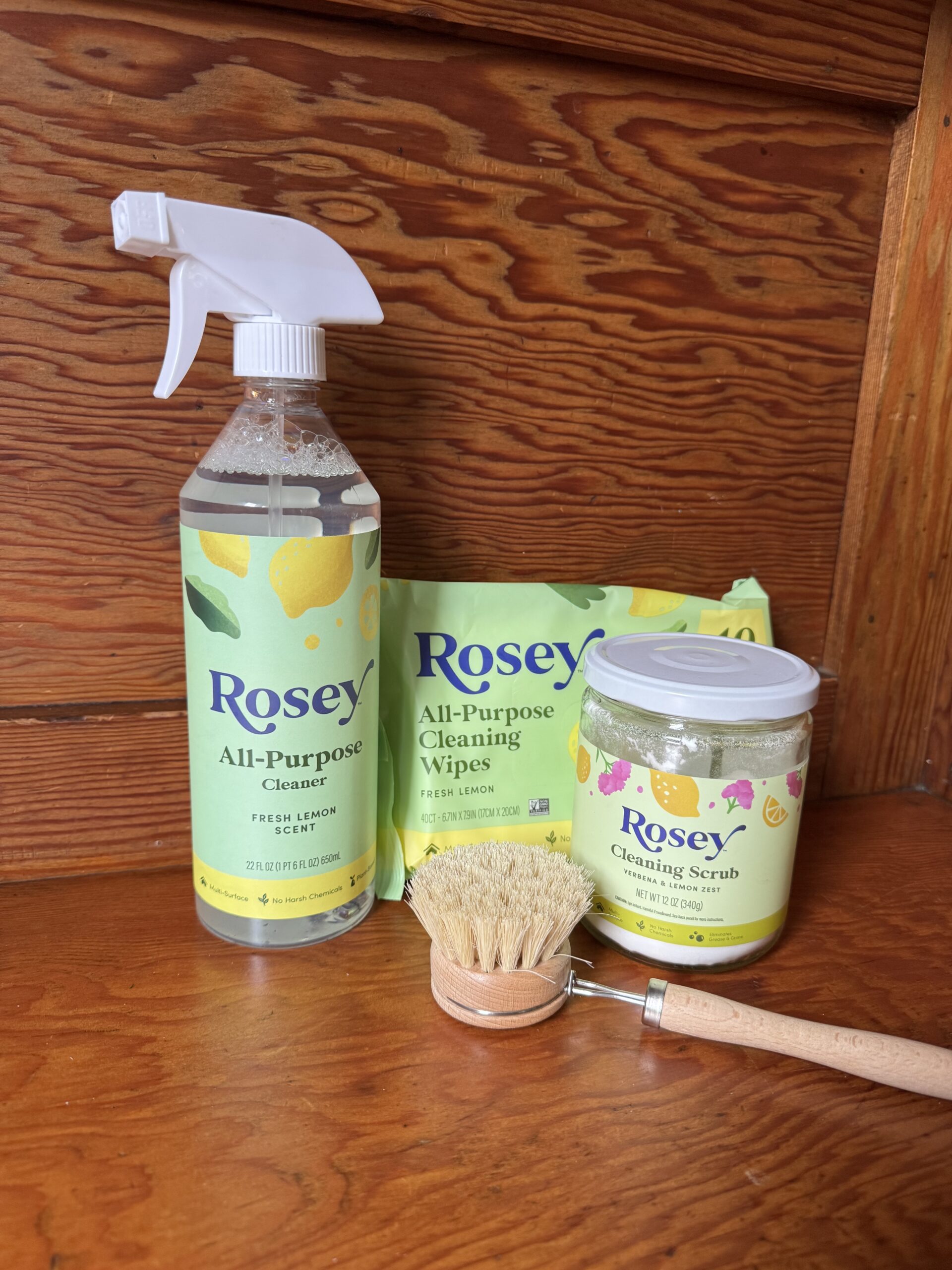 A bottle of Rosey all-purpose cleaner, a pack of cleaning wipes, a jar of cleaning scrub, and a wooden scrub brush on a wood surface.