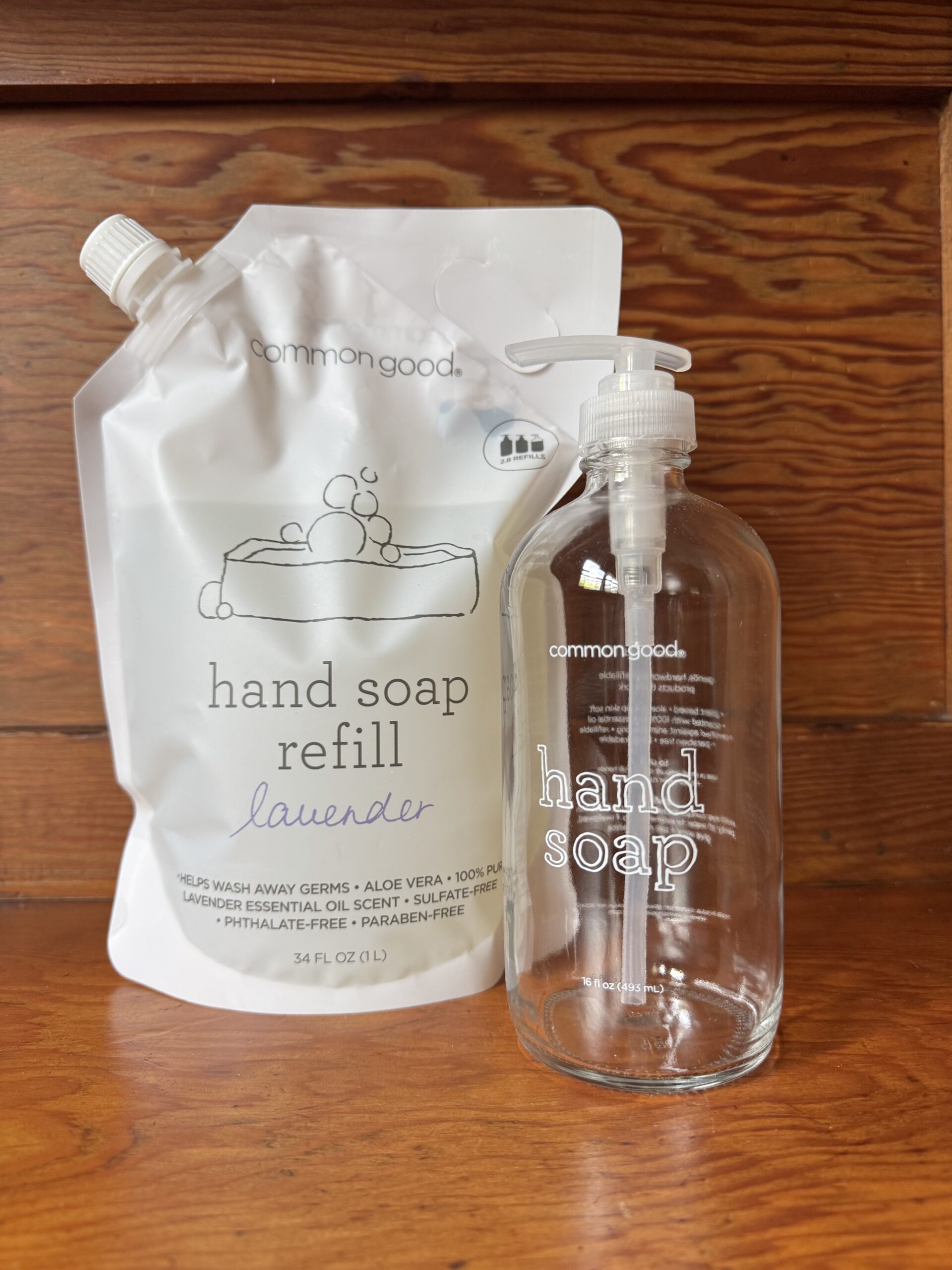 A lavender hand soap refill pouch and an empty glass hand soap dispenser with a pump are placed side by side on a wooden surface.
